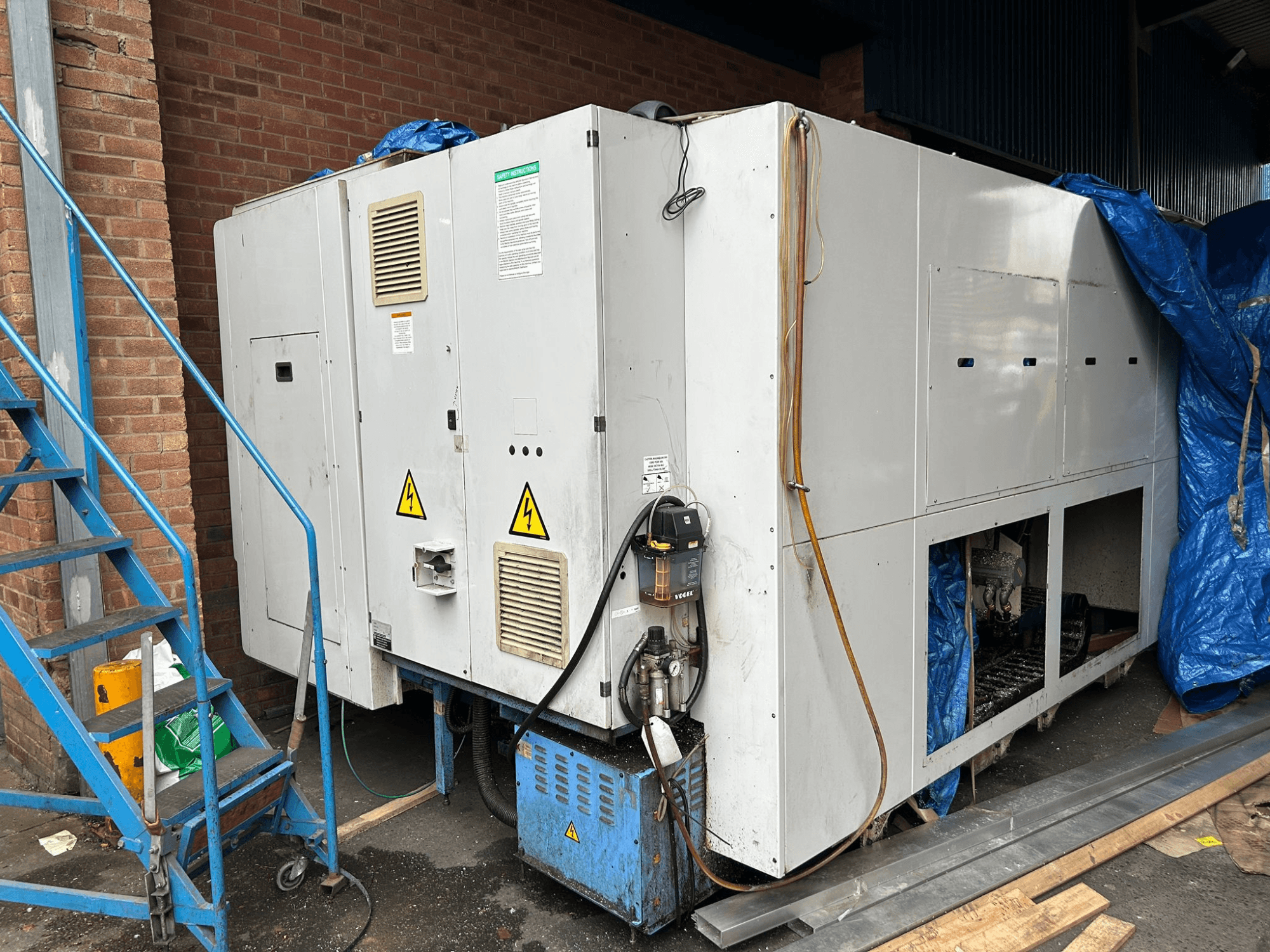 Egy nagyméretű ipari hűtőberendezés oldalnézetből, márka és modell nem tisztázott, elektromos csatlakozók, figyelmeztető táblák és kék ponyvával fedett.