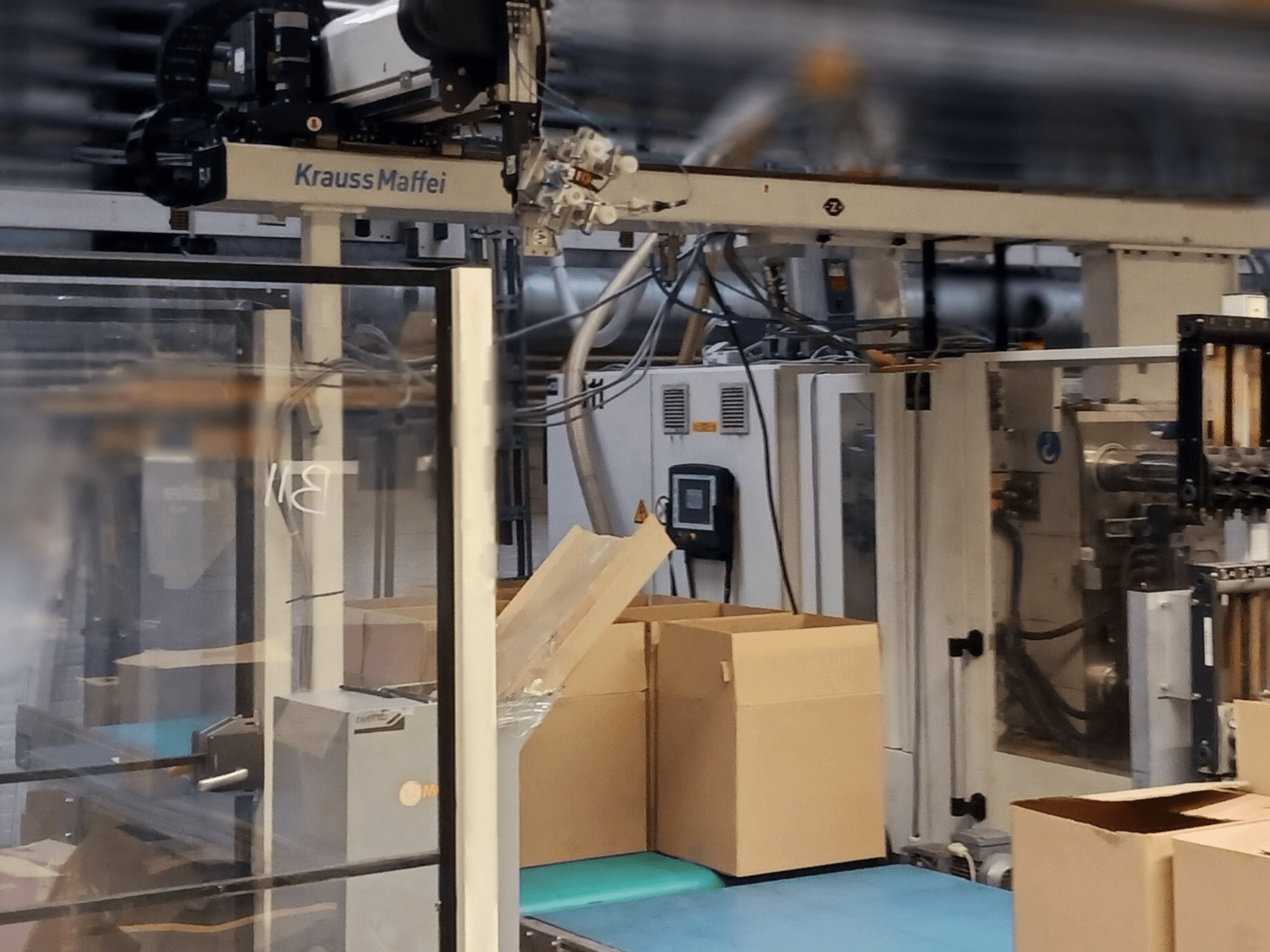 Krauss Maffei automatizálási rendszer egy gyárban, elölnézetben a szállítószalagon lévő dobozok és a gépelemek működés közben.