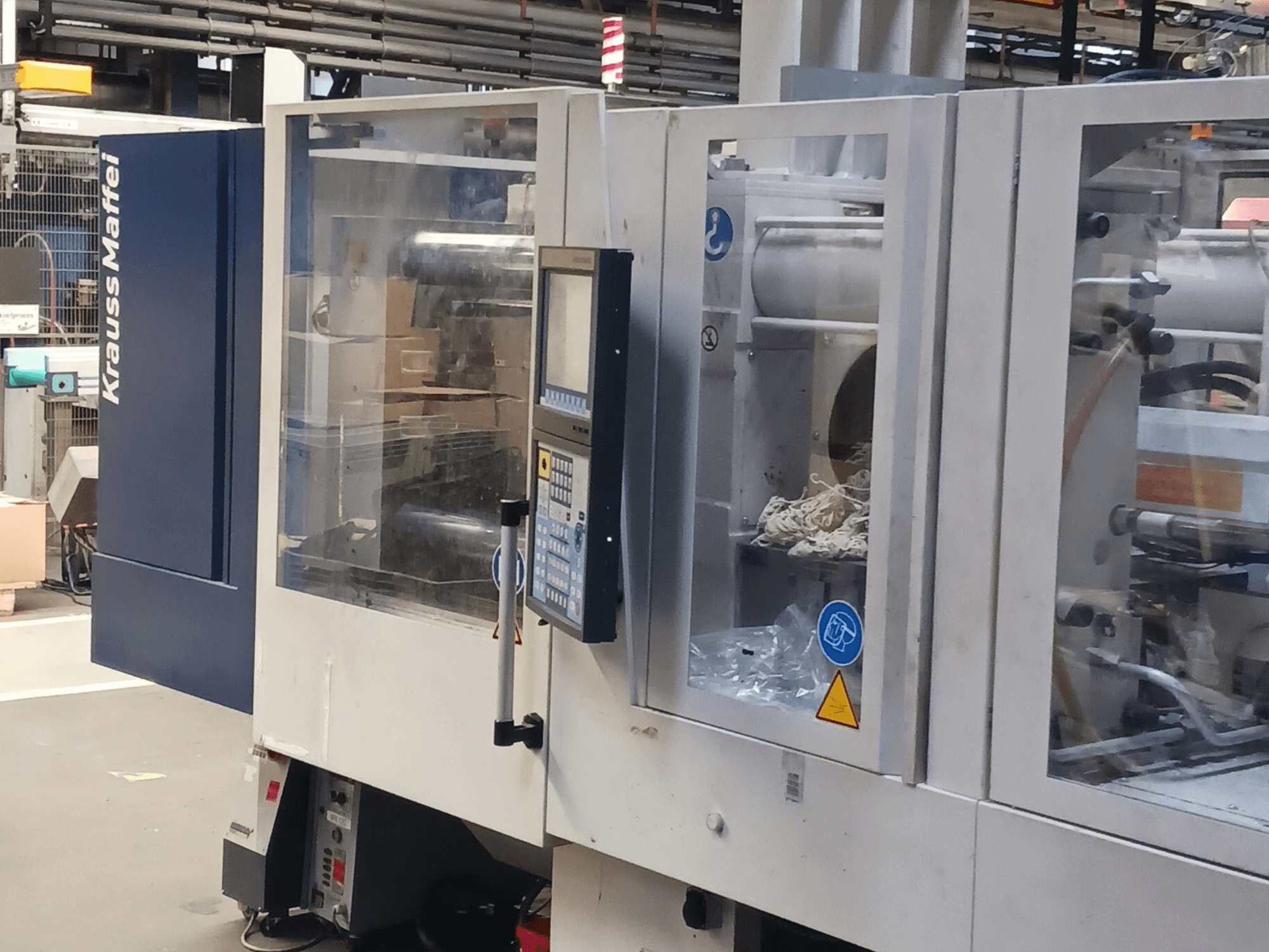 KraussMaffei fröccsöntőgép, oldalnézet, a vezérlőpanel, a gépek és a csomagolóanyagok a háttérben.