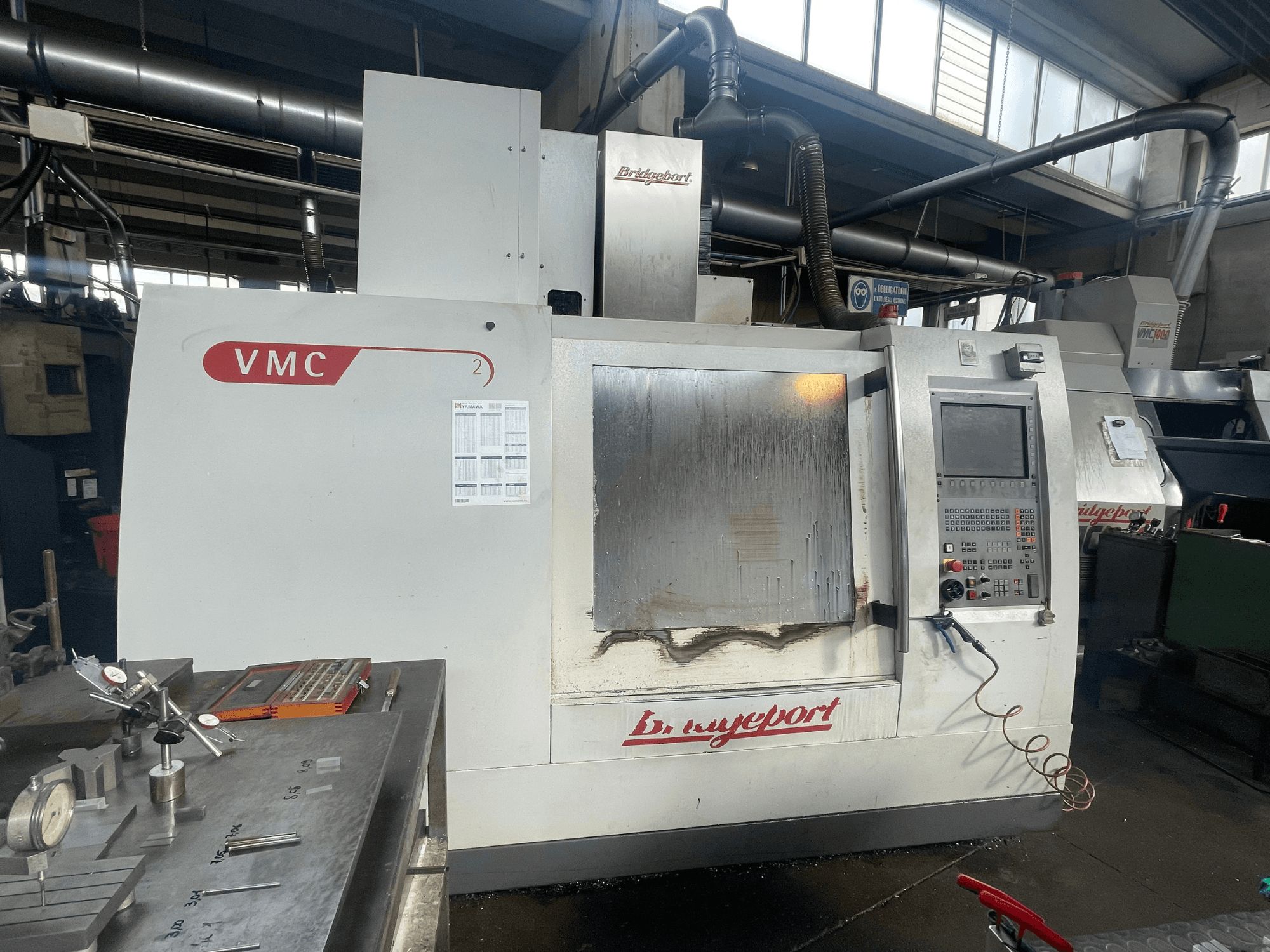 A Bridgeport VCM 800-30 függőleges megmunkáló központ egy műhelyben, vezérlőpanellel és szerszámtáblával. Elölnézet, látható részletekkel.