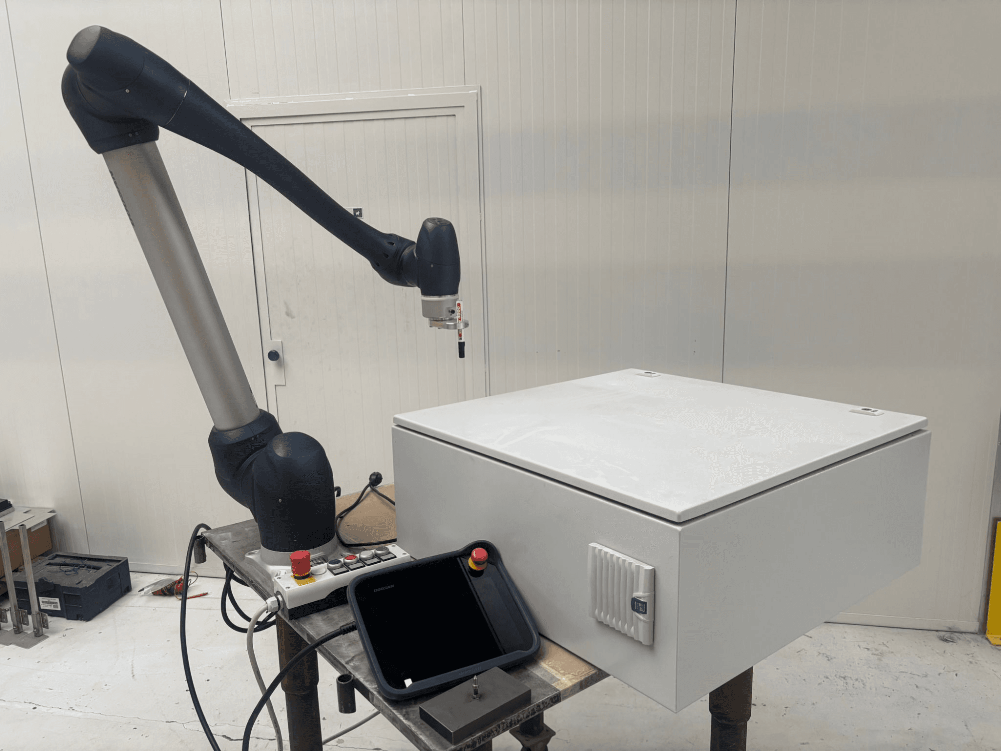 DOOSAN M0617 kollaboratív robot oldalnézetben, a robotkar kinyújtva a vezérlőfelület és egy asztalon lévő fehér doboz fölött.