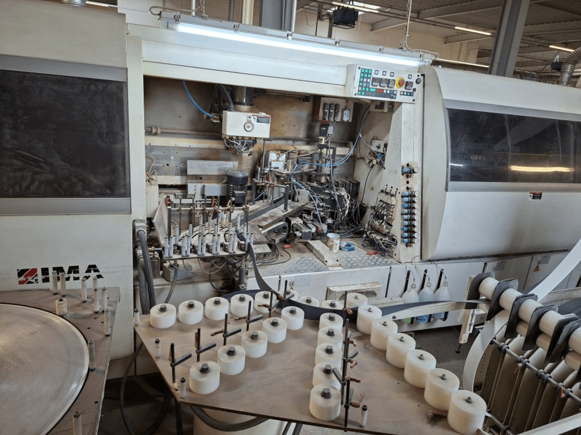 IMA gépek oldalról nézve, összetett mechanikai alkatrészek és egy kerek asztal fehér orsókkal.