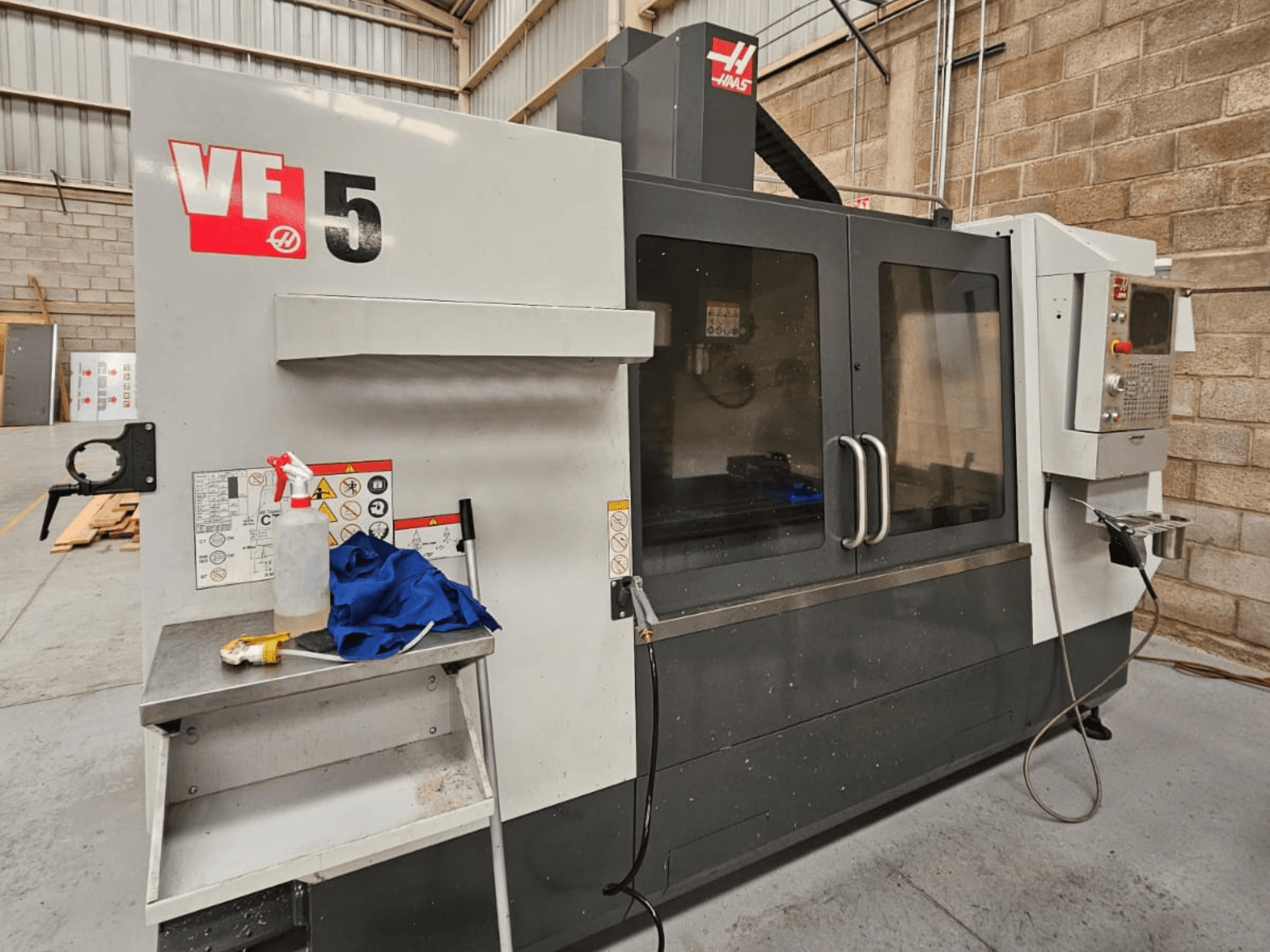 Haas VF-5 CNC marógép elölnézetben, kezelőpanellel, hűtőfolyadékos palackkal, kék ruhával és biztonsági utasításokkal.