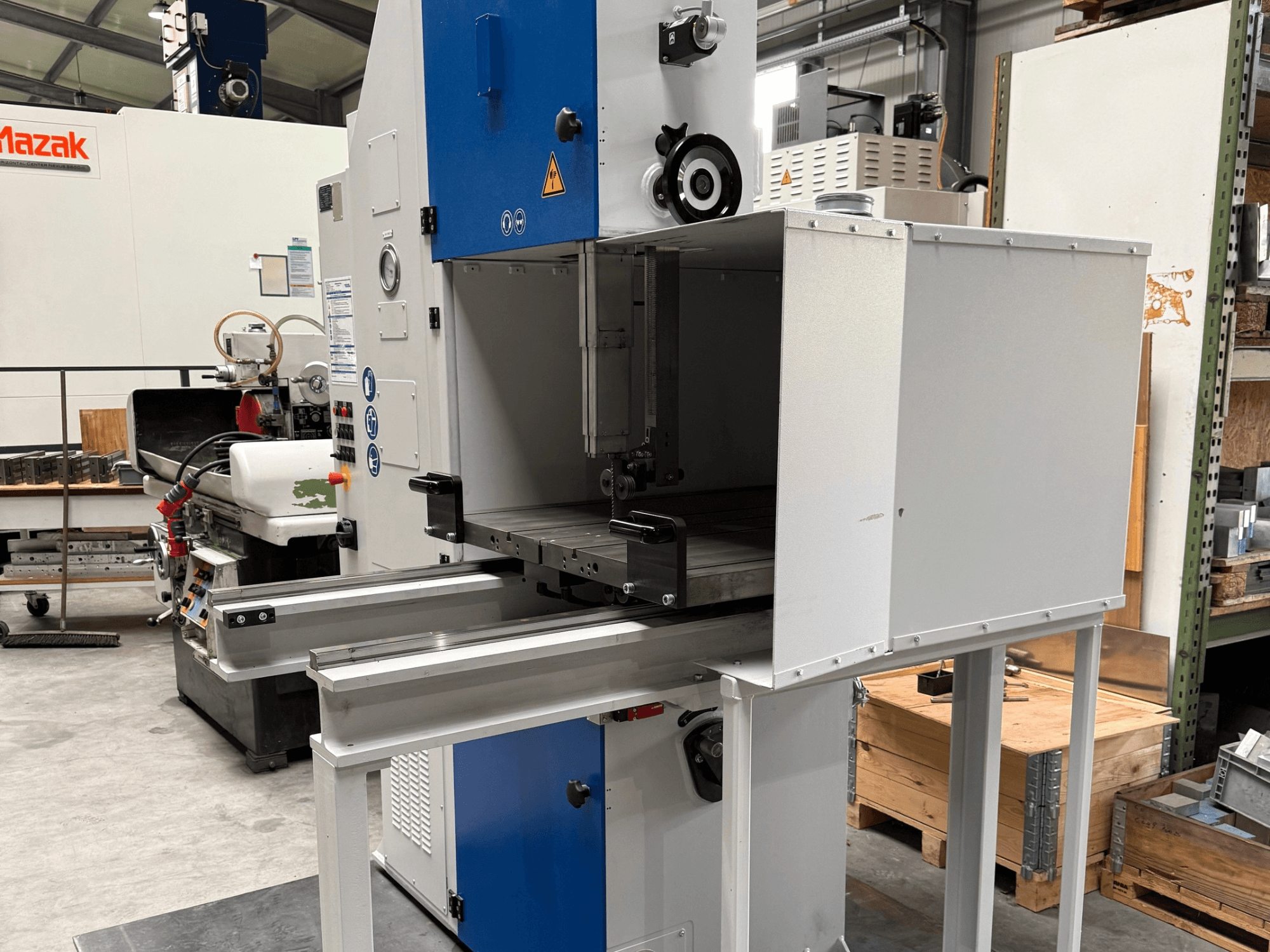 Mazak ipari gép szemből nézve, kék vezérlőpanellel, fém asztallal és biztonsági figyelmeztetésekkel.