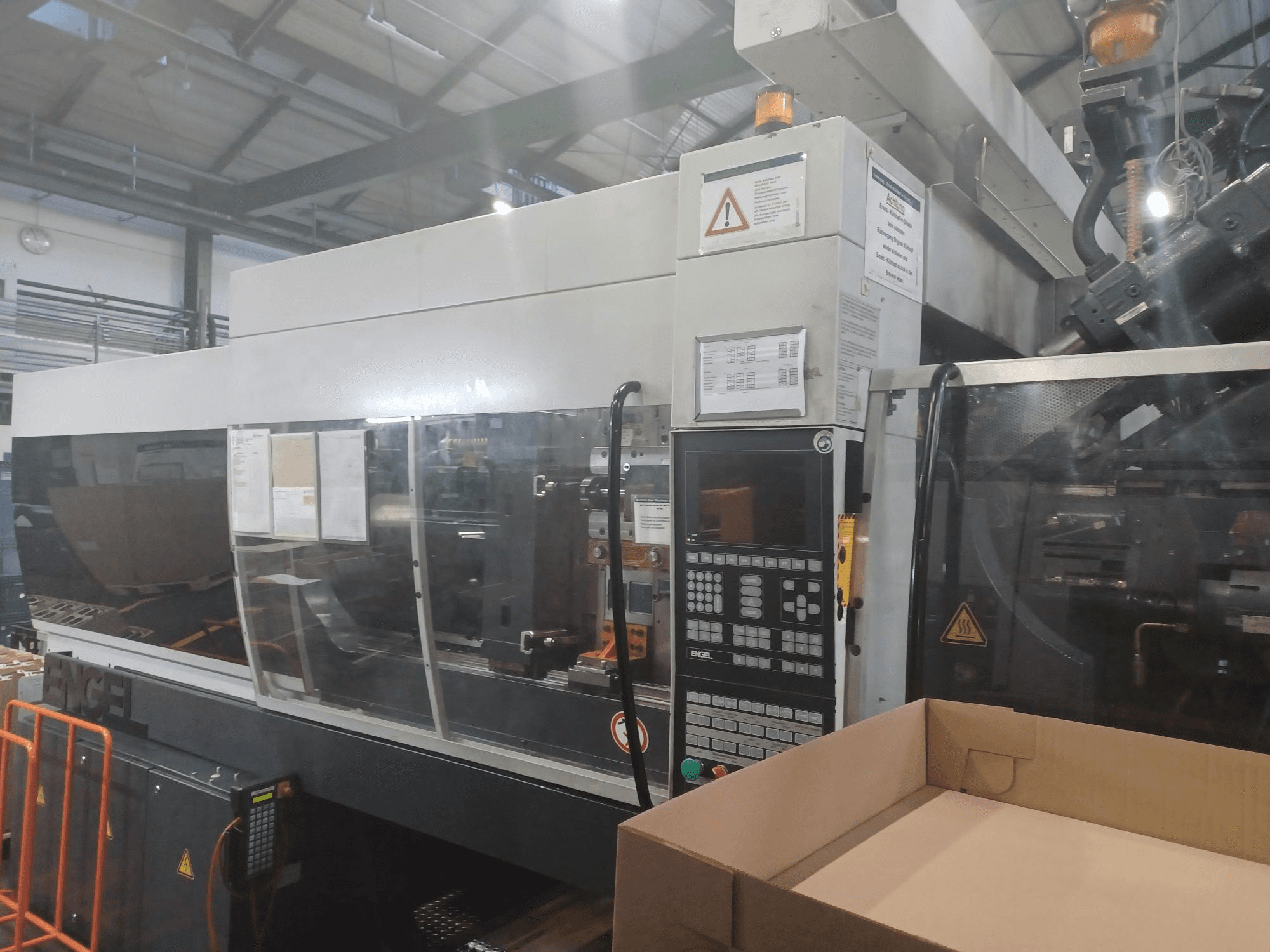 Engel fröccsöntőgép, elölnézet, vezérlőpanel, biztonsági címkék és egy üres doboz a közelben.
