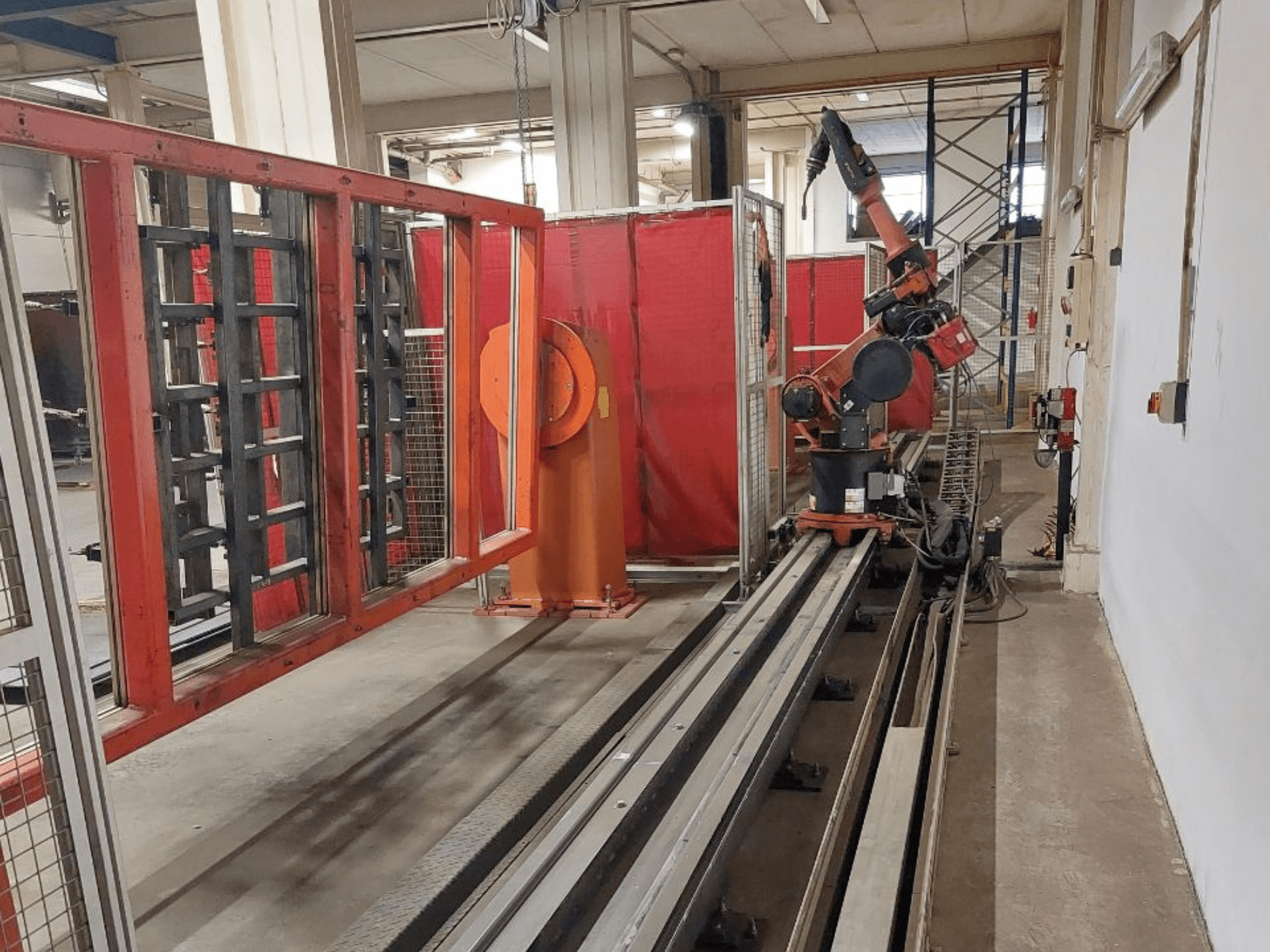 Narancssárga és fekete színű ipari robotkar fémpályákon, piros védőfalakkal körülvéve, elölnézetben.