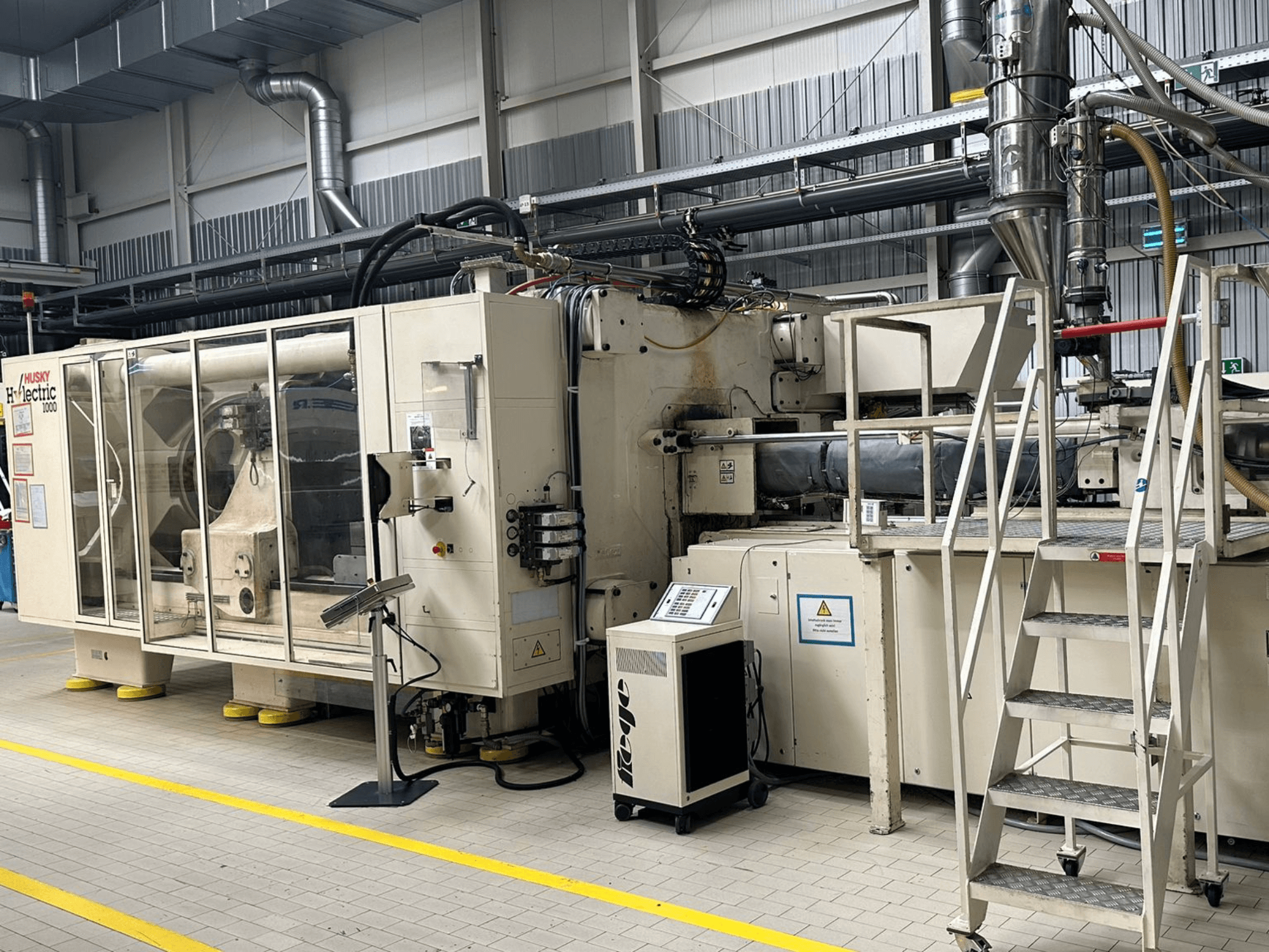 A Husky Hylelectric H1000 RS95/80 elektromos fröccsöntőgép elölnézete gyári környezetben, a vezérlőpanellel és a lépcsőkkel együtt.