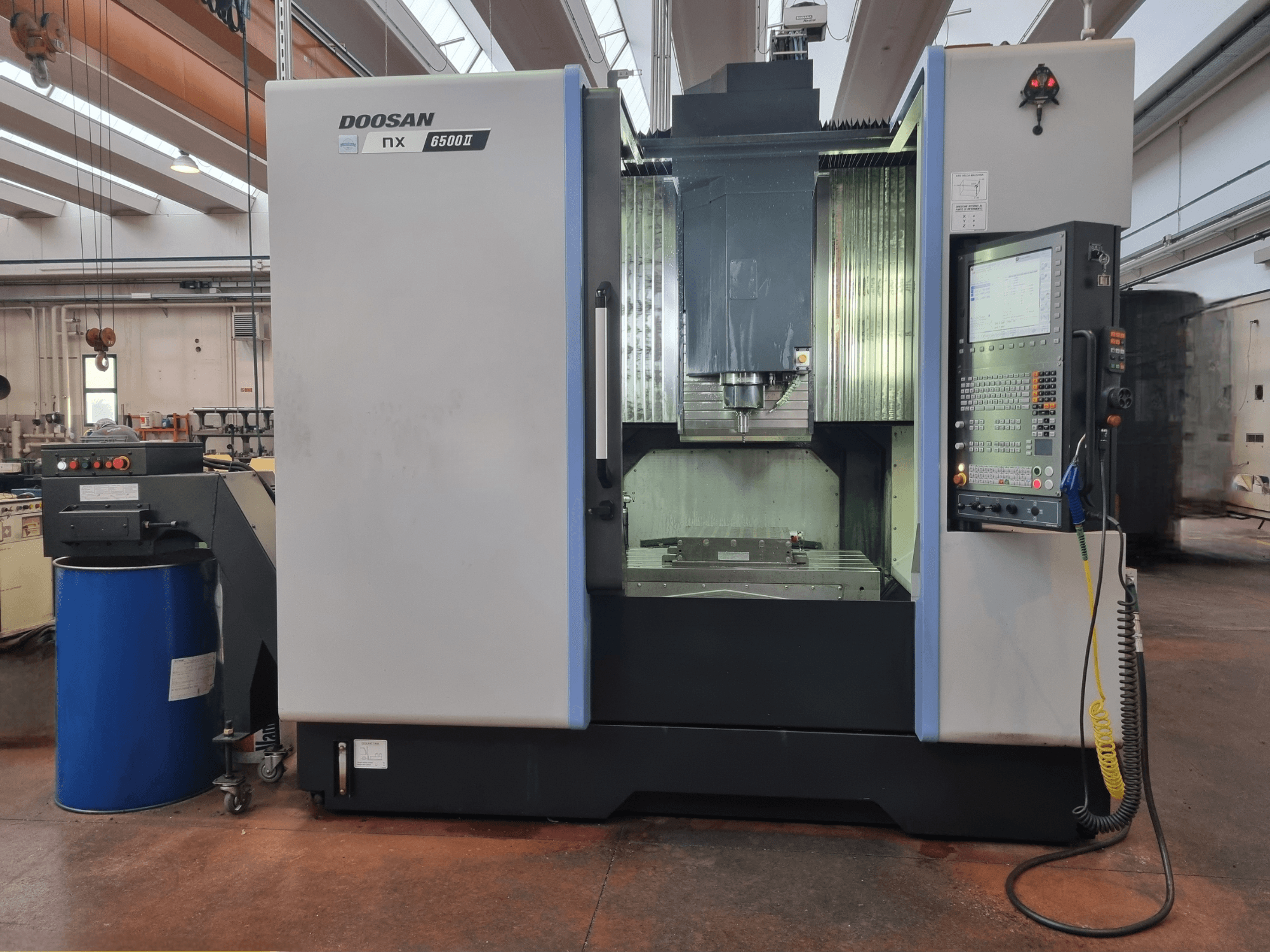 Doosan NX 6500 II CNC-gép, előlnézet, a vezérlőpanel és a megmunkálóterület, egy kék hordó mellett.