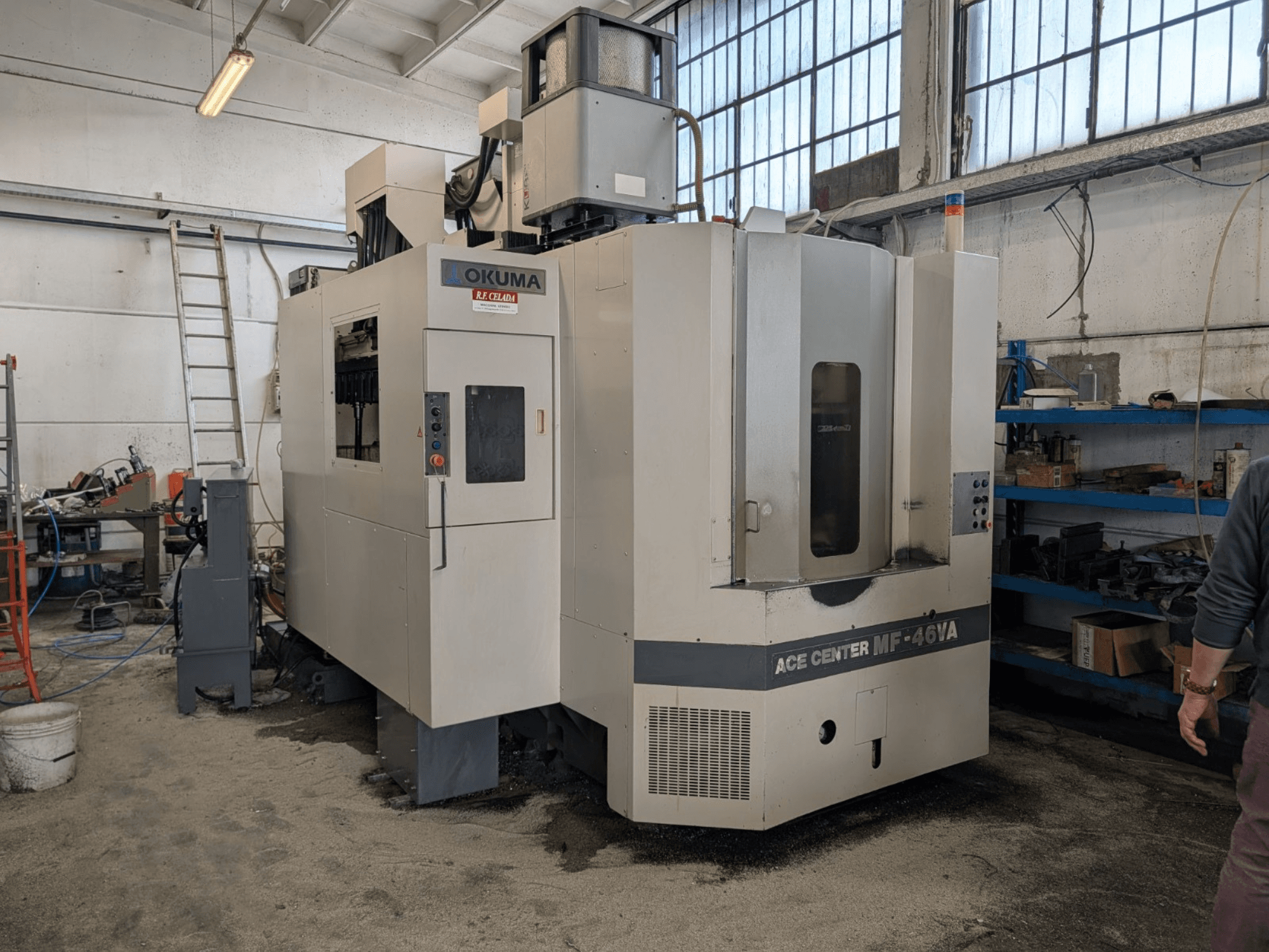 Okuma ACE CENTER MF-46VA függőleges megmunkálóközpont egy műhelyben, elölnézetben a vezérlőpanellel és a környező berendezésekkel.