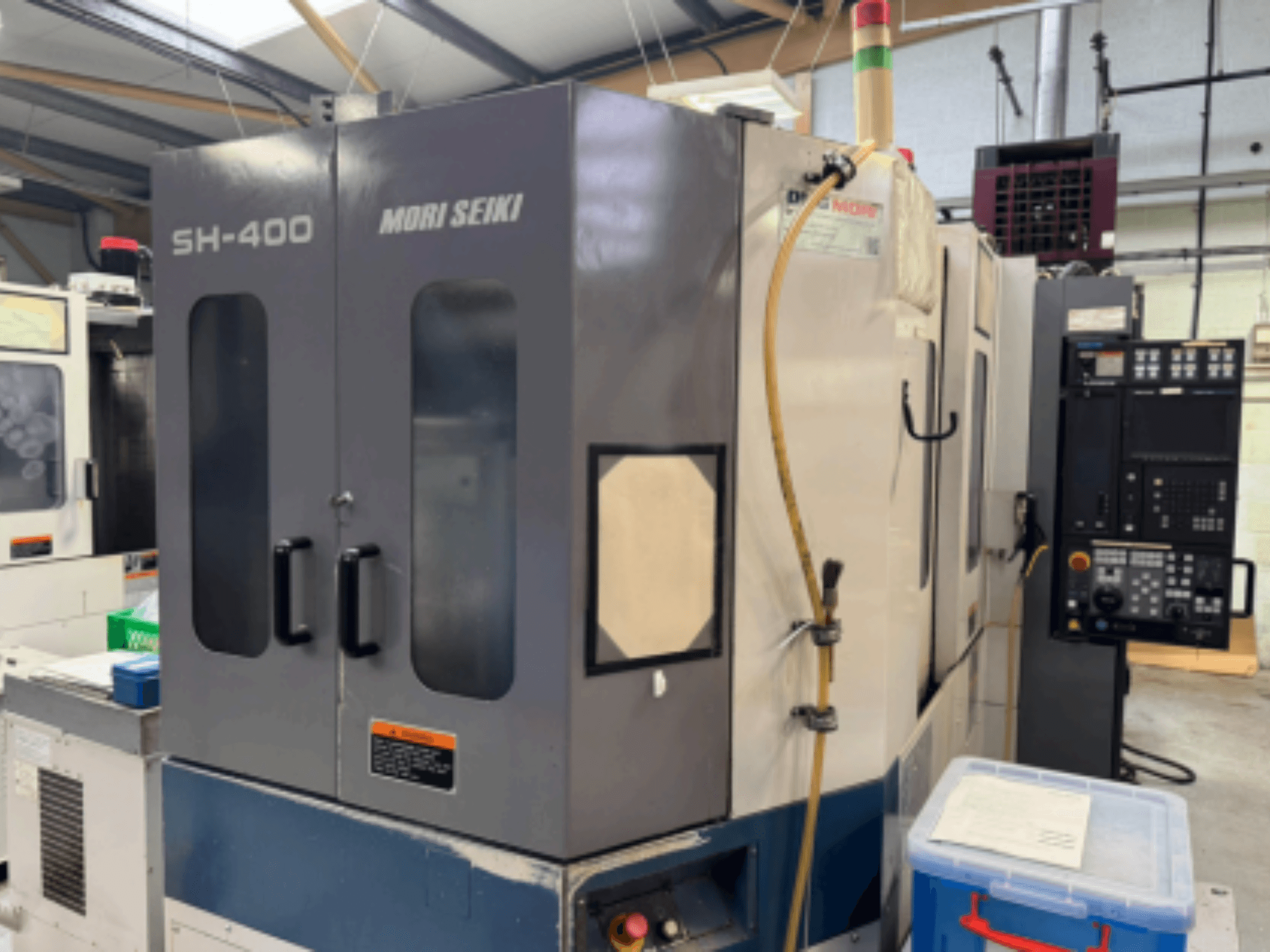 Mori Seiki SH-400 CNC gép, elölnézet, kettős ajtó, vezérlőpanel és rögzítőtömlő. A közelben kék tároló konténer.