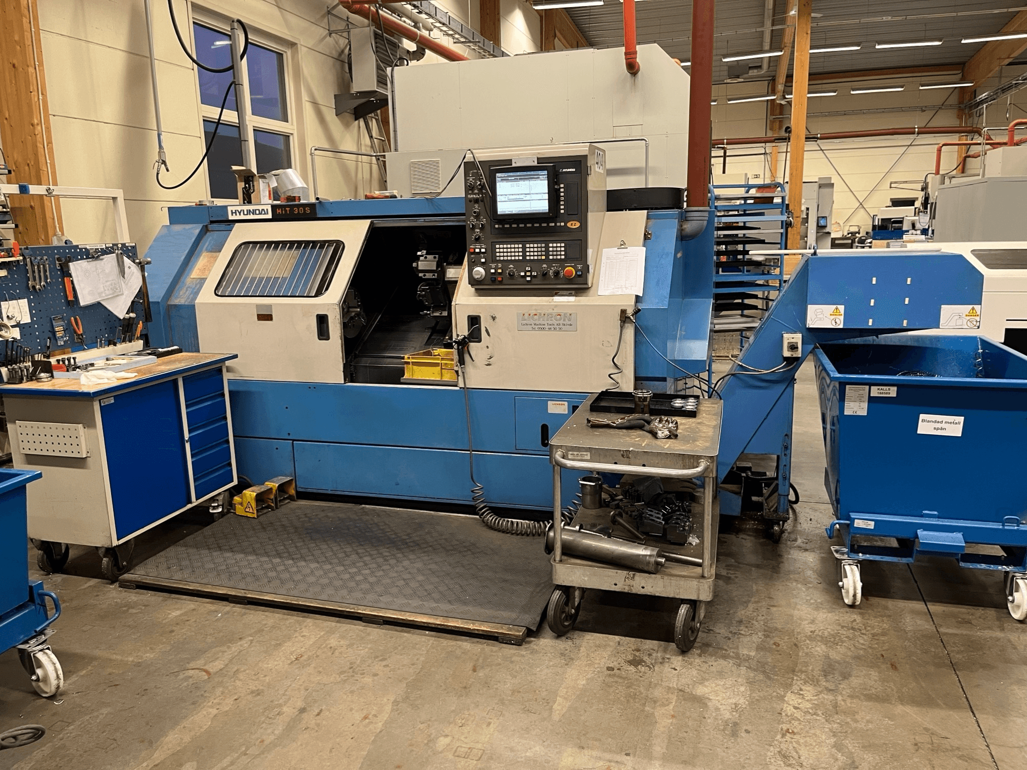 Hyundai HIT 30S vízszintes esztergagép, elölnézetben, kezelőpanellel és szerszámokkal egy közeli kocsin, műhelyben.