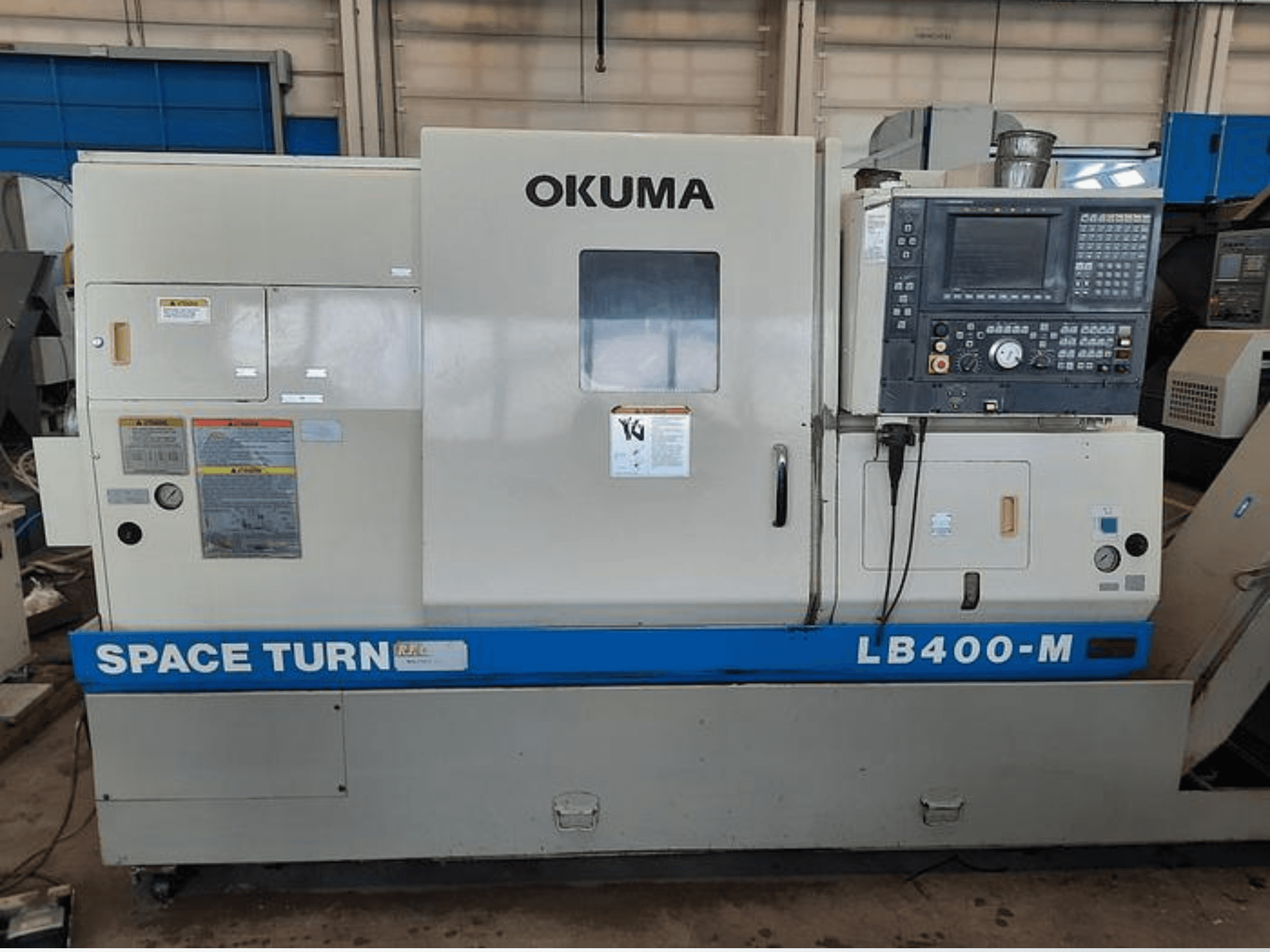 Okuma LB400-M CNC esztergagép, elölnézet a vezérlőpanellel, a figyelmeztető címkékkel és a kék "SPACE TURN" márkajelzéssel.