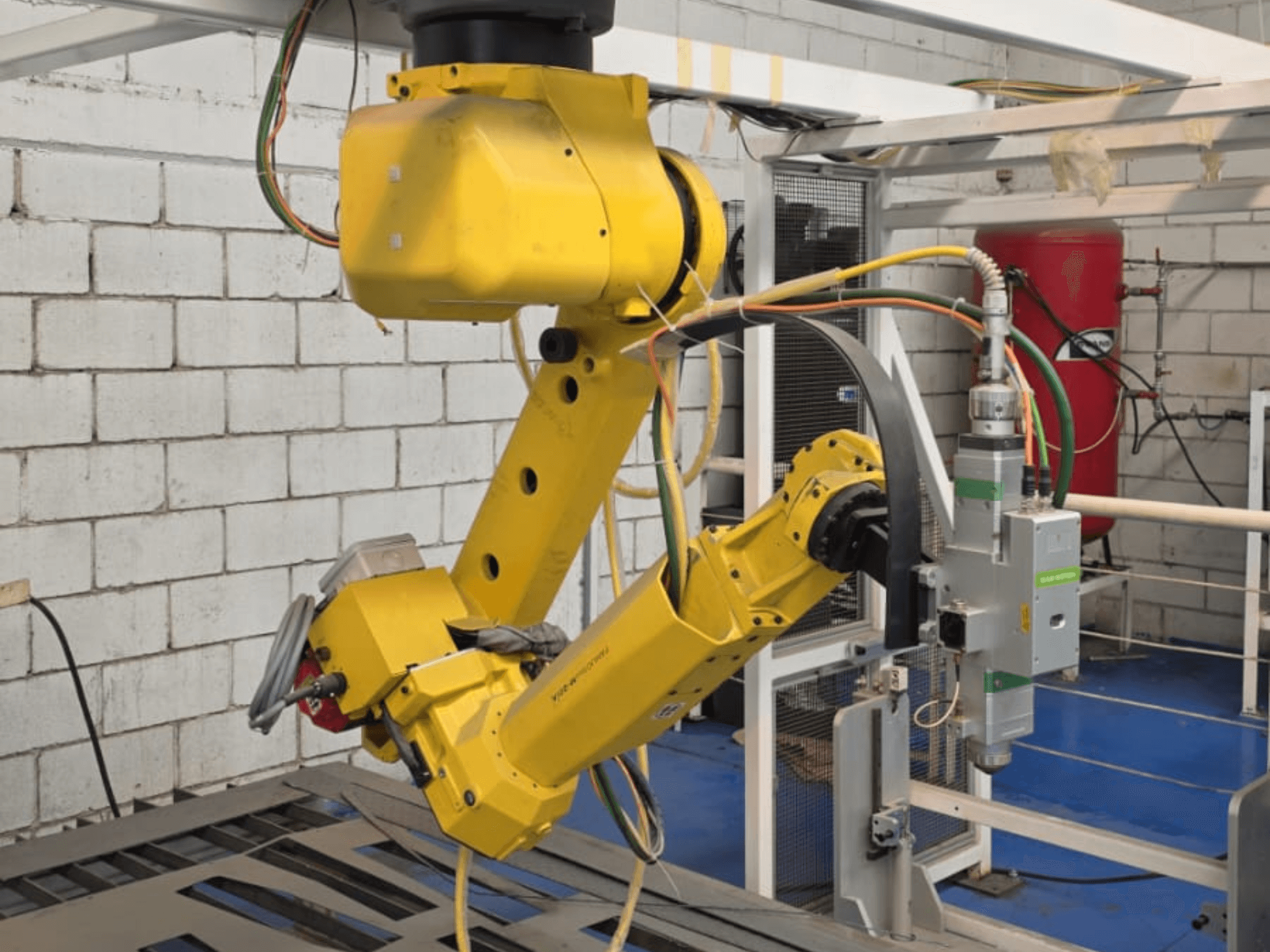Sárga robotkar egy precíziós szerszámmal, több ízülettel és kábellel, szemből nézve, ipari környezetben.