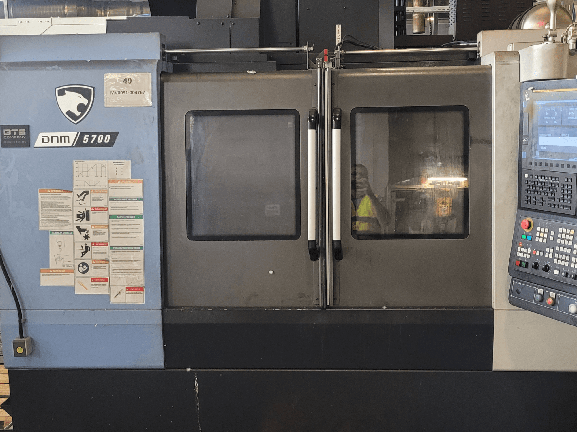 A DOOSAN DNM 5700 függőleges megmunkáló központ elölnézete a vezérlőpanellel, a biztonsági címkékkel és a kissé résnyire nyitott ajtókkal.