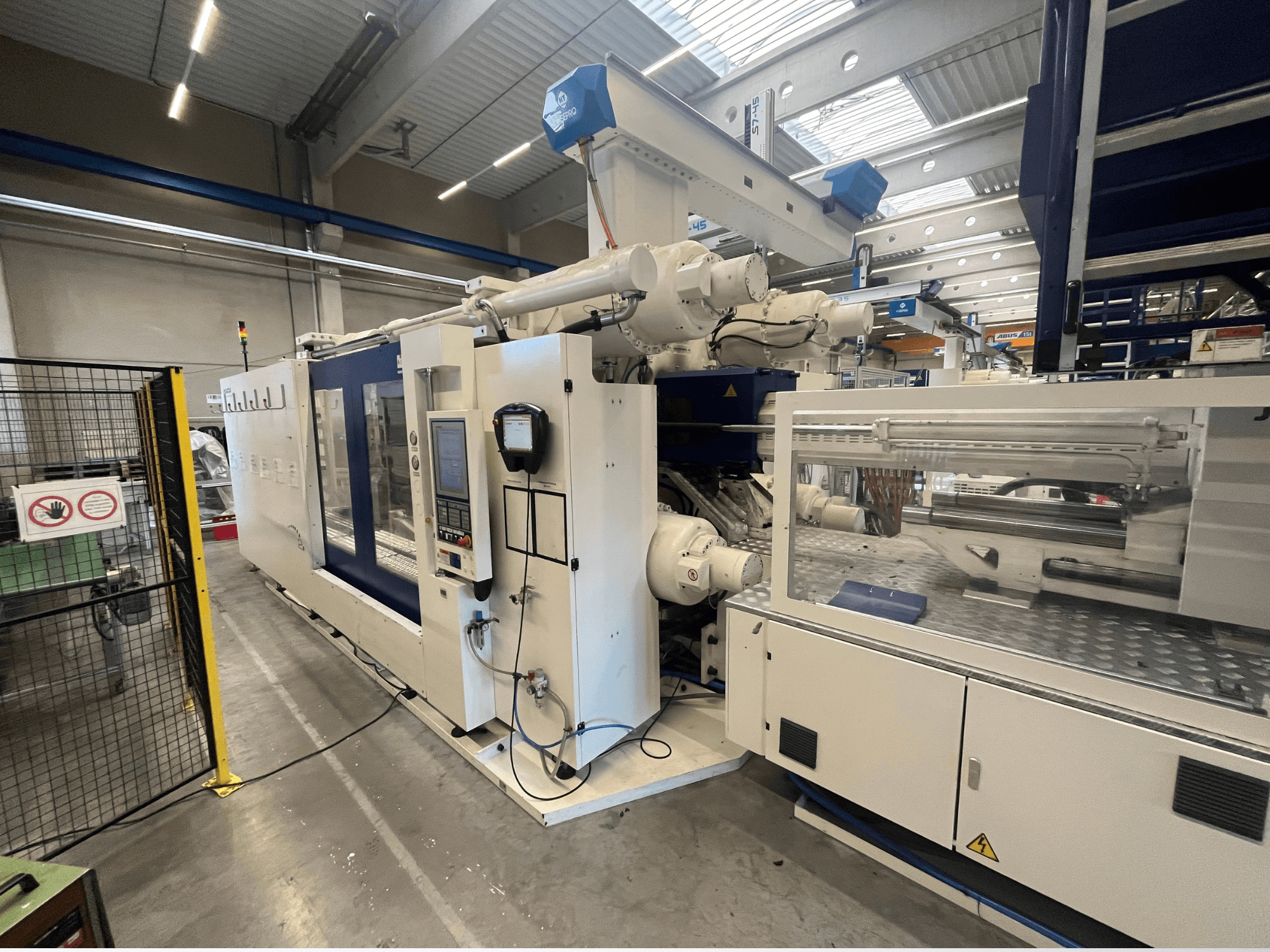 Fehér és kék színű fröccsöntőgép, elölnézetben, vezérlőpanellel, robotkarral és biztonsági jelzésekkel.