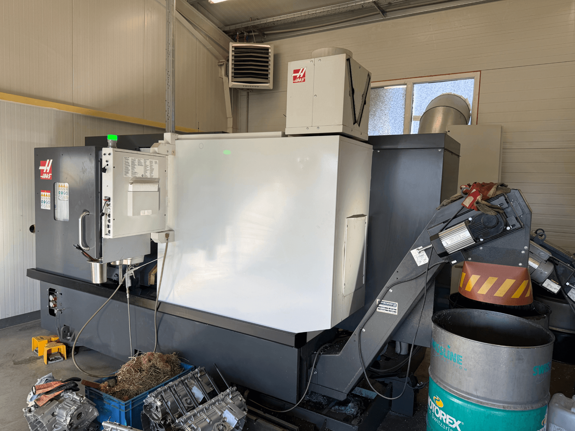 Haas CNC-gép vezérlőpanellel, oldalnézet; látható hűtőfolyadék- és olajvezetékek; fémhulladék a kék kukában; ipari környezet.