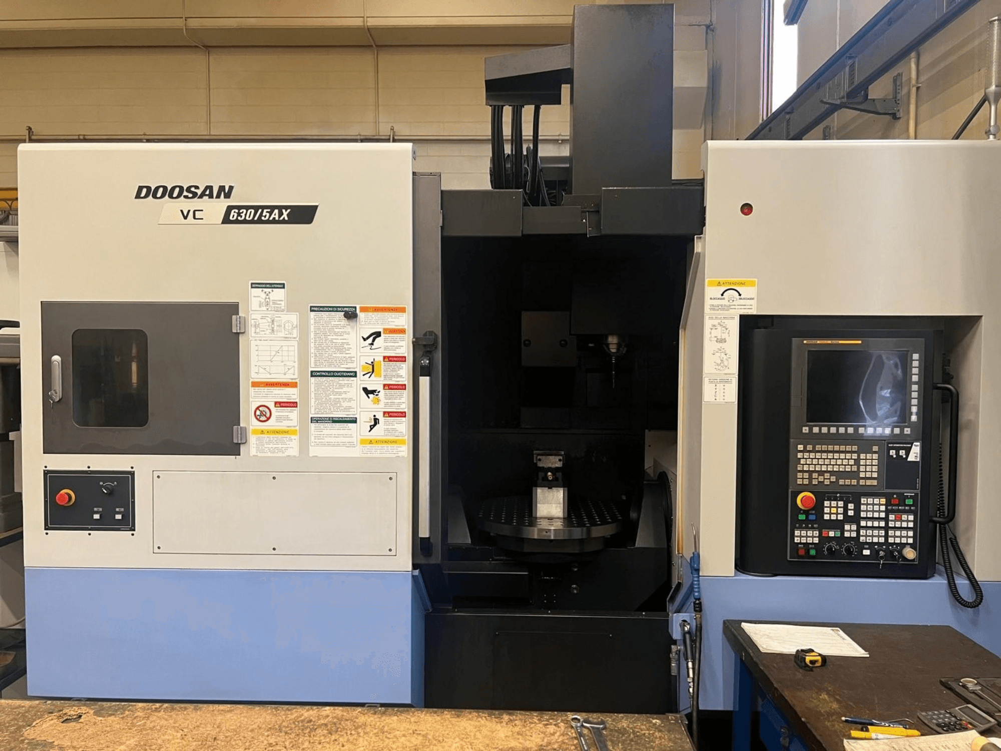 A DOOSAN VC 630/5AX függőleges megmunkáló központ elölnézete, amelyen látható a vezérlőpanel és az orsóterület, valamint a biztonsági címkék.