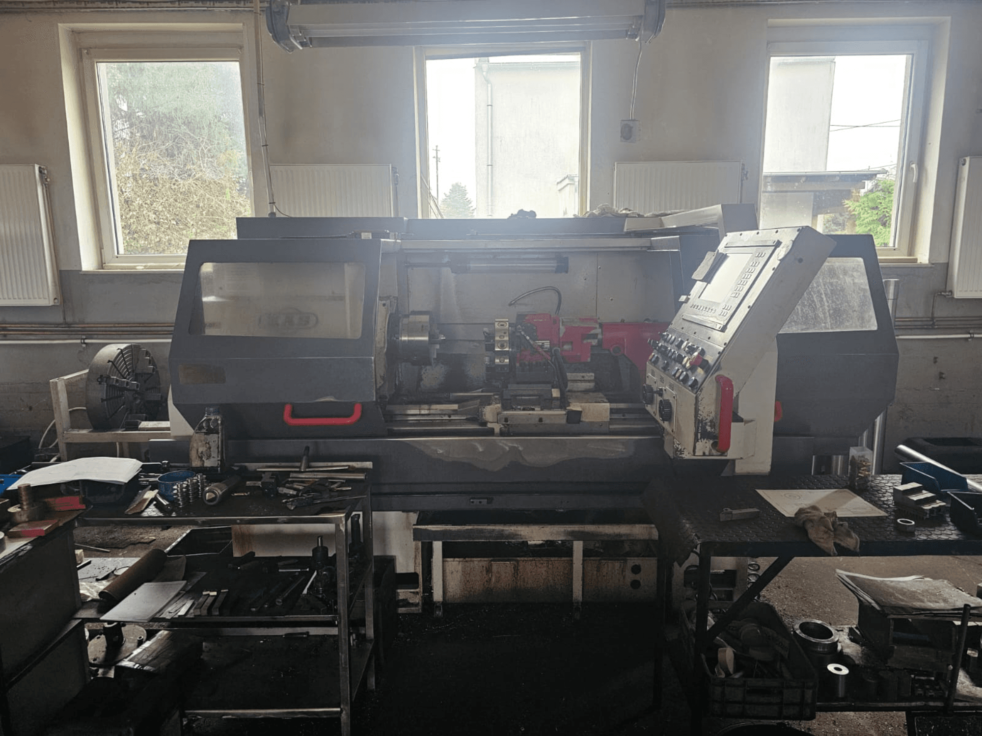 Esztergagép egy műhelyben, elölnézetben, vezérlőpanellel és szerszámokkal az asztalokon, ablakokkal és rendetlen munkaterülettel körülvéve.