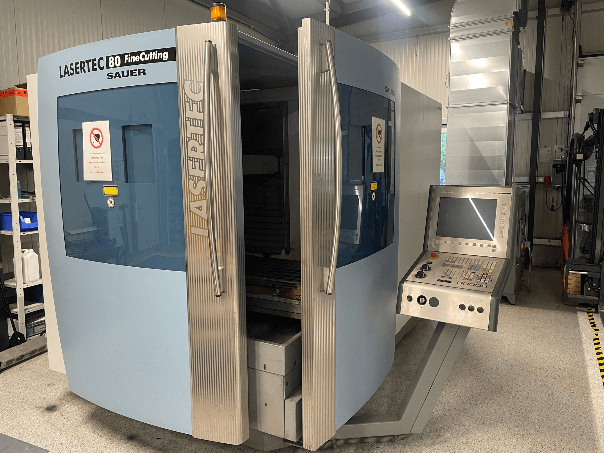 A DMG Sauer Lasertec80 FineCutting lemezvágó gép elölnézete a vezérlőpanellel, nyitott ajtókkal és látható biztonsági jelzésekkel.