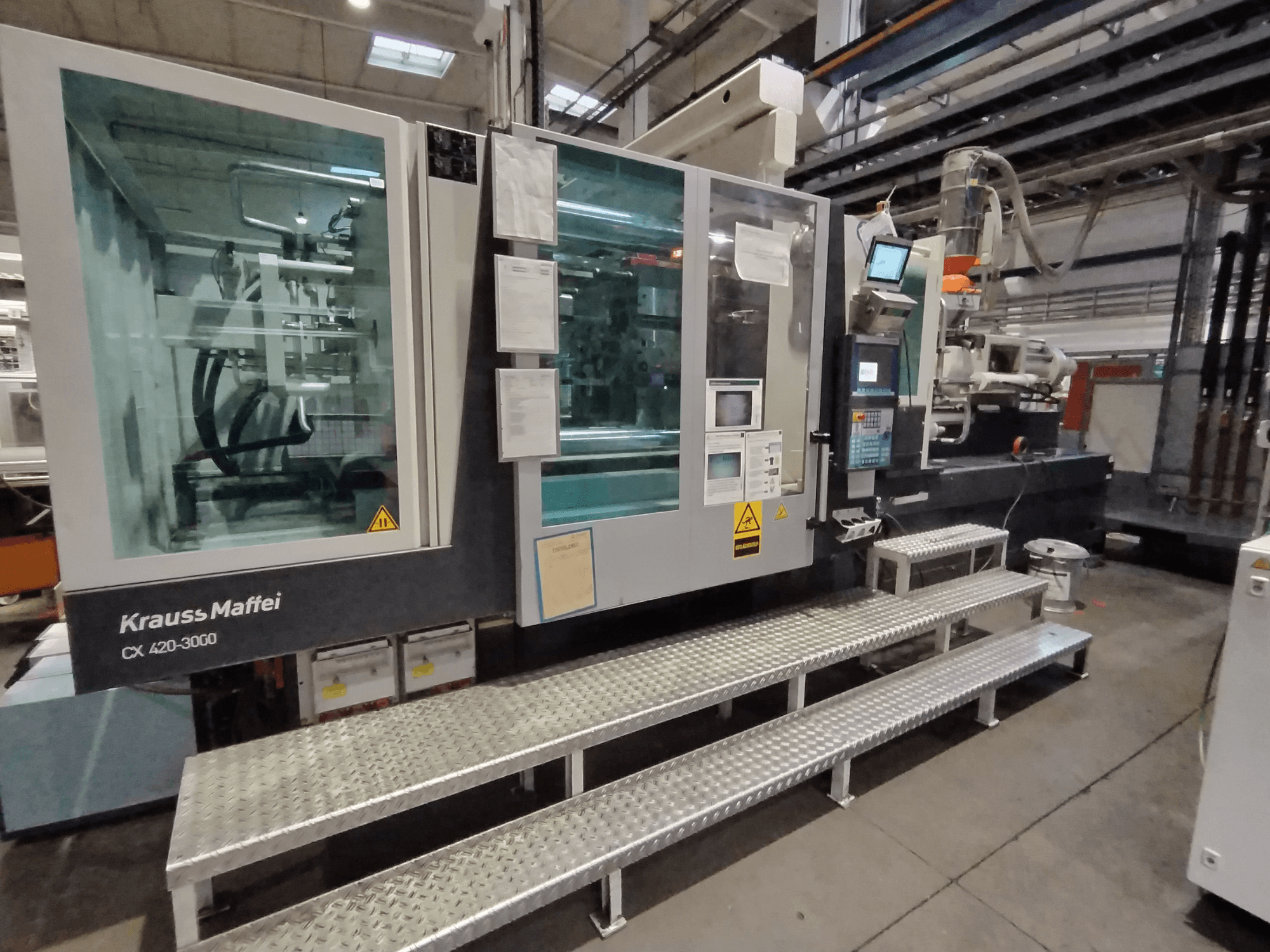 Krauss Maffei CX 420-3000 fröccsöntőgép, oldalnézet, látható vezérlőpanel és biztonsági jelzések, fémlépcsőn állítva.
