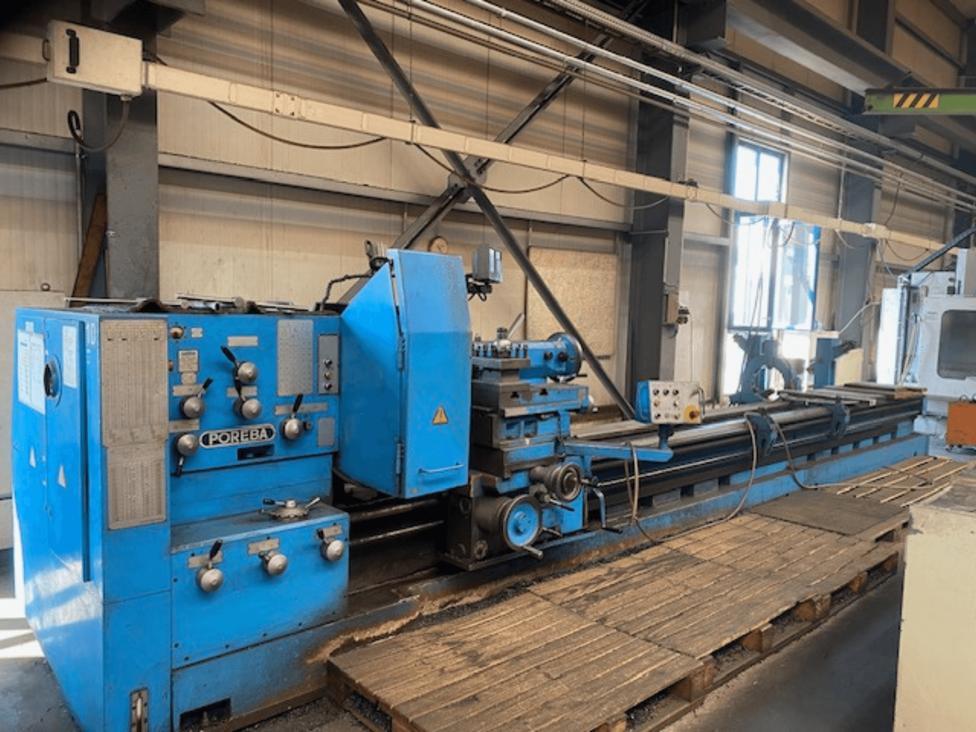 Egy kék színű POREBA esztergagép elülső nézete, vezérlőgombokkal, szerszámtámasszal és alatta lévő fa raklapokkal.