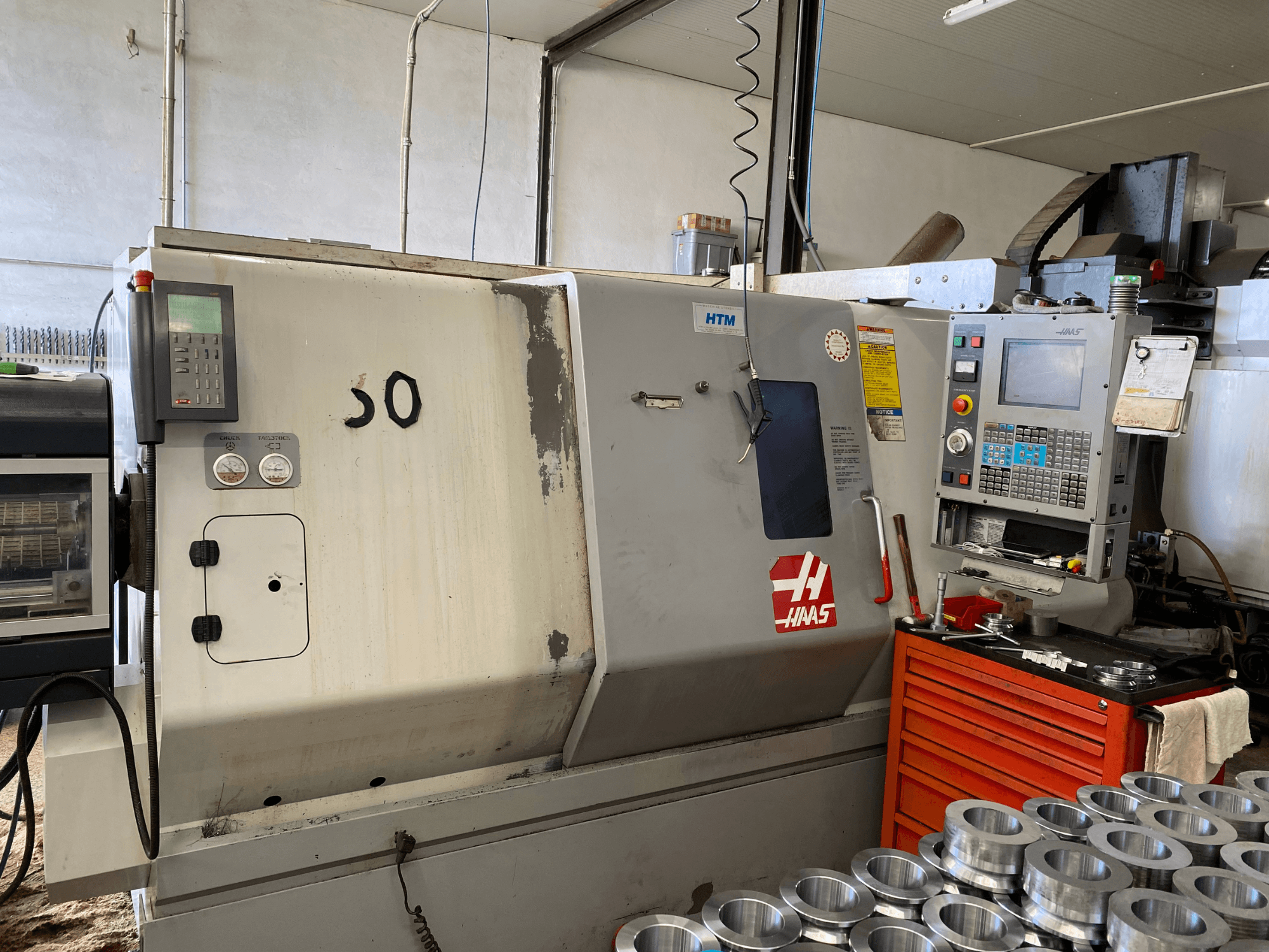 Haas CNC-gép vezérlőpanellel, elölnézetben, a kezelőgombok, a műszerek és a közelben elhelyezett fém alkatrészek.