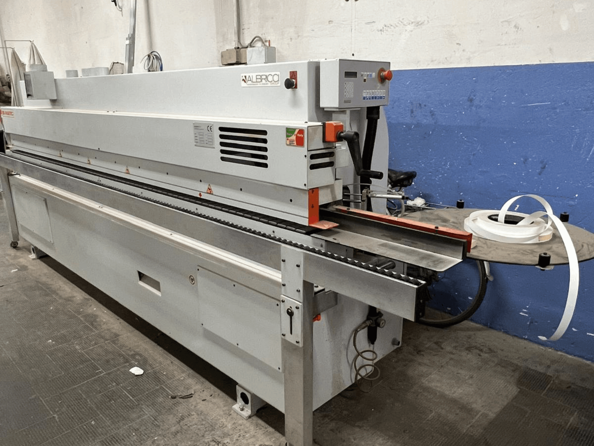 ALBRICO élszalagozó gép elölnézetben, hosszú szállítószalaggal és egy csatlakoztatott ragasztószalag-tekerccsel.