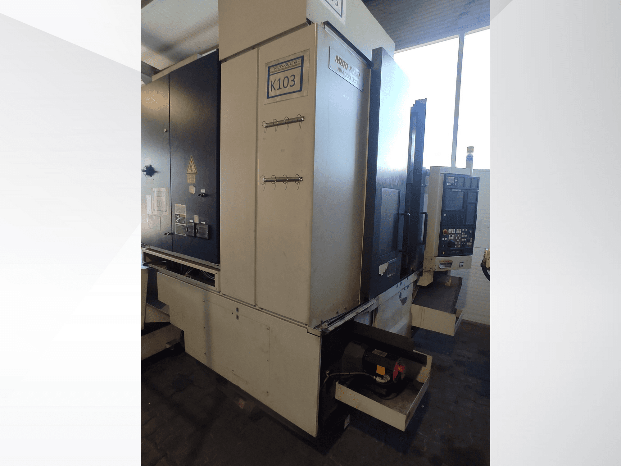 Mori Seiki K103 CNC-gép elölnézetben, a vezérlőpanel, a biztonsági címkék és az elektromos alkatrészek láthatóak.
