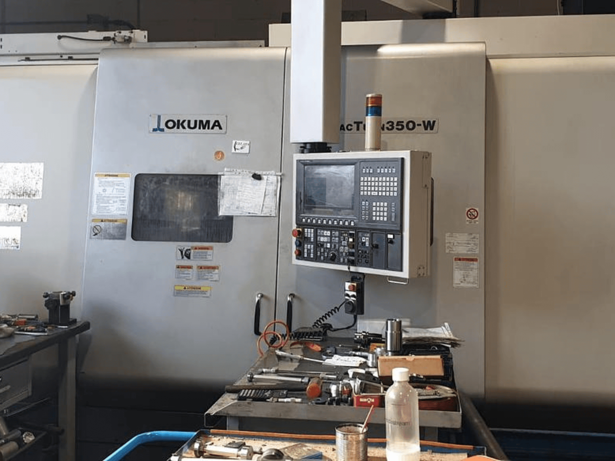 Okuma MAC TURN 350-W esztergafúró központ, elölnézetben a vezérlőpanellel és a szerszámokkal és egy palackkal zsúfolt munkaterülettel.