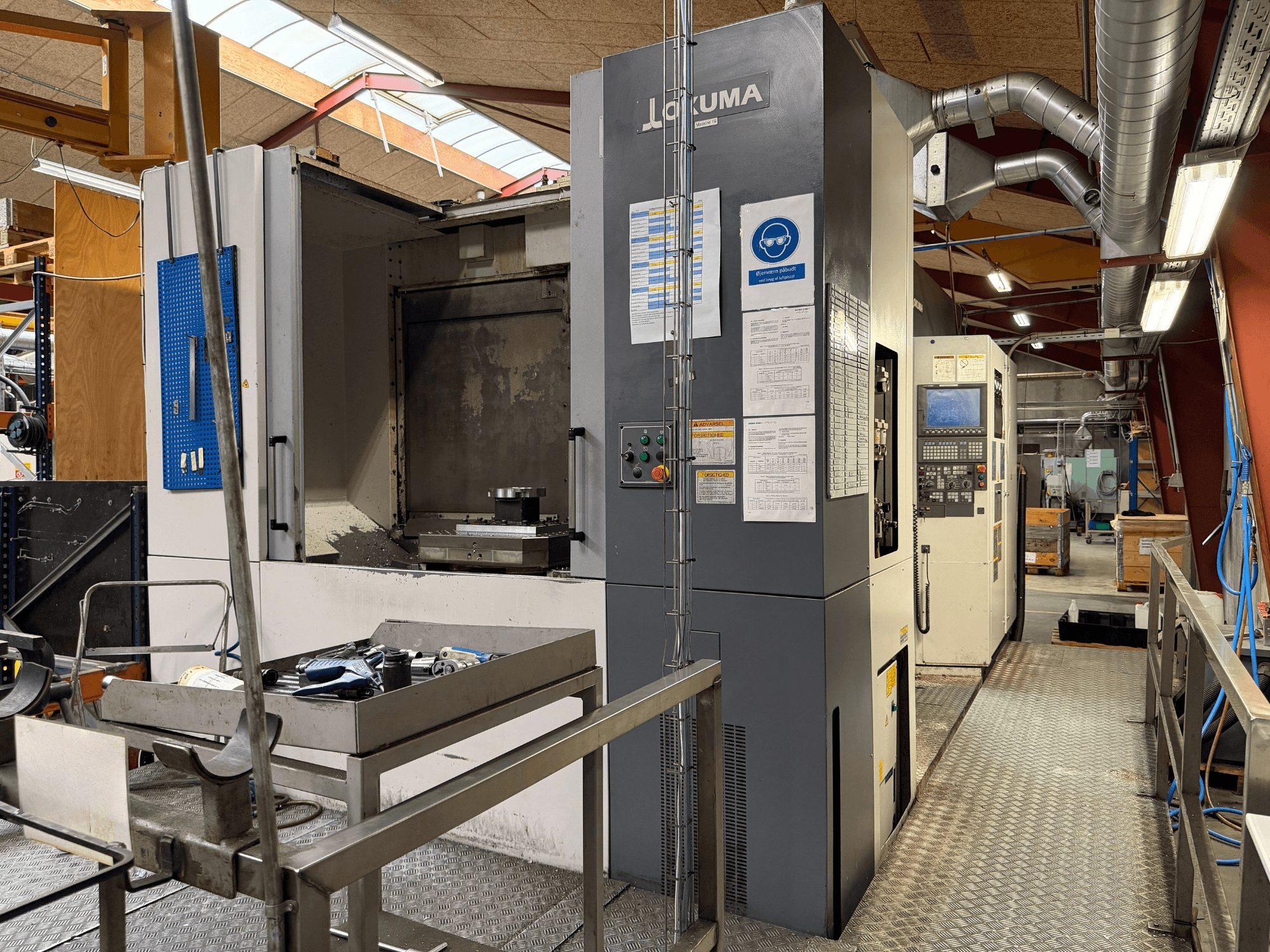Okuma MA-600HB vízszintes megmunkáló központ, elölnézet, amelyen láthatók a fémasztalon elhelyezett szerszámok és a gép vezérlőelemei.