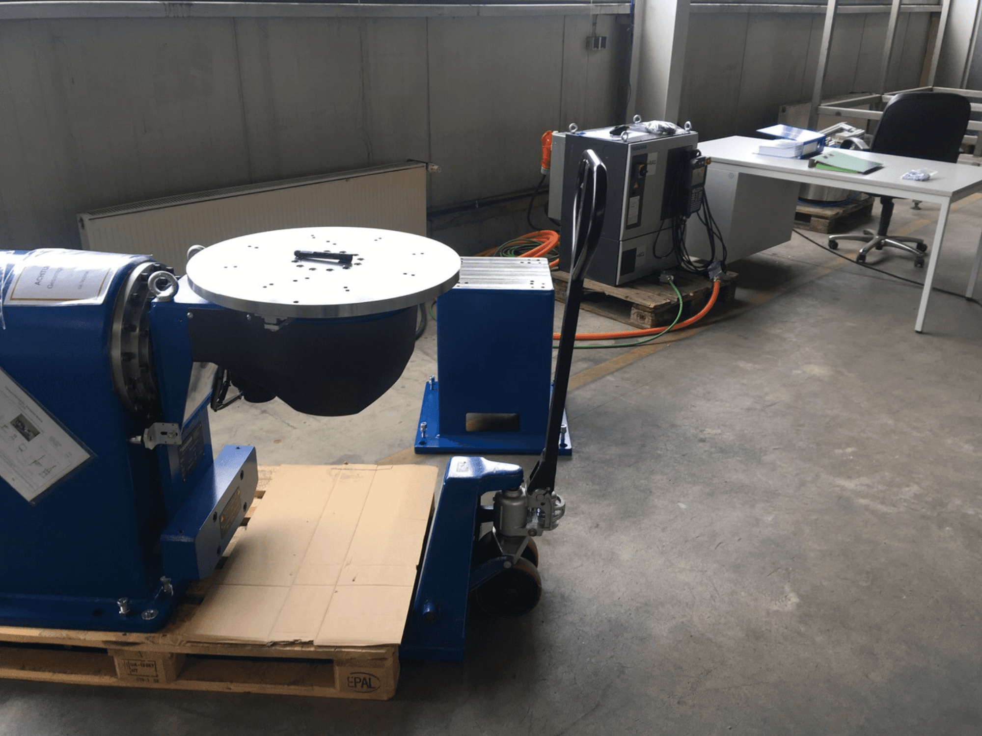 Kék ipari berendezés kör alakú talapzattal és vezérlőpanellel, elölről nézve, fából készült raklapon, irodai környezetben.