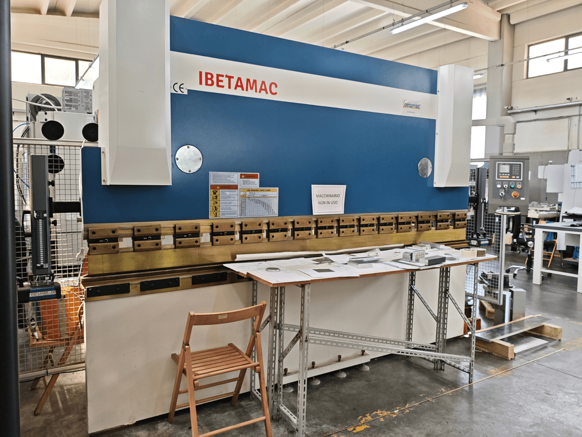 IBETAMAC 3200 100T hajlítógép egy műhelyben, oldalnézetből, amelyen látható a vezérlőpanel, a munkaterület és a biztonsági figyelmeztetések.