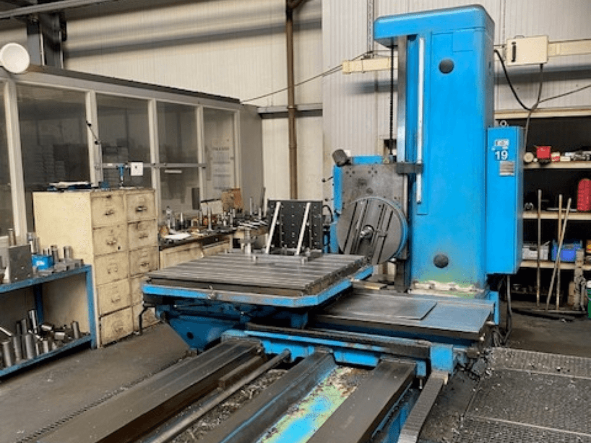 Kék marógép elölnézetben, 19-es modellszámú, műhelypadlón, a háttérben szerszámok és egy tárolószekrény.