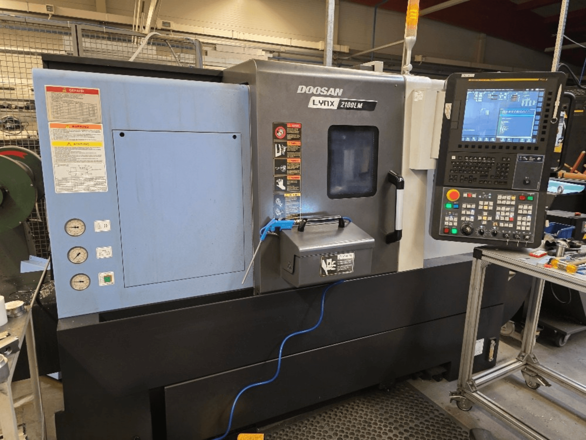 Doosan Lynx 2100LM CNC eszterga, előlnézet, vezérlőpanel, nyomásmérők és biztonsági figyelmeztetések.
