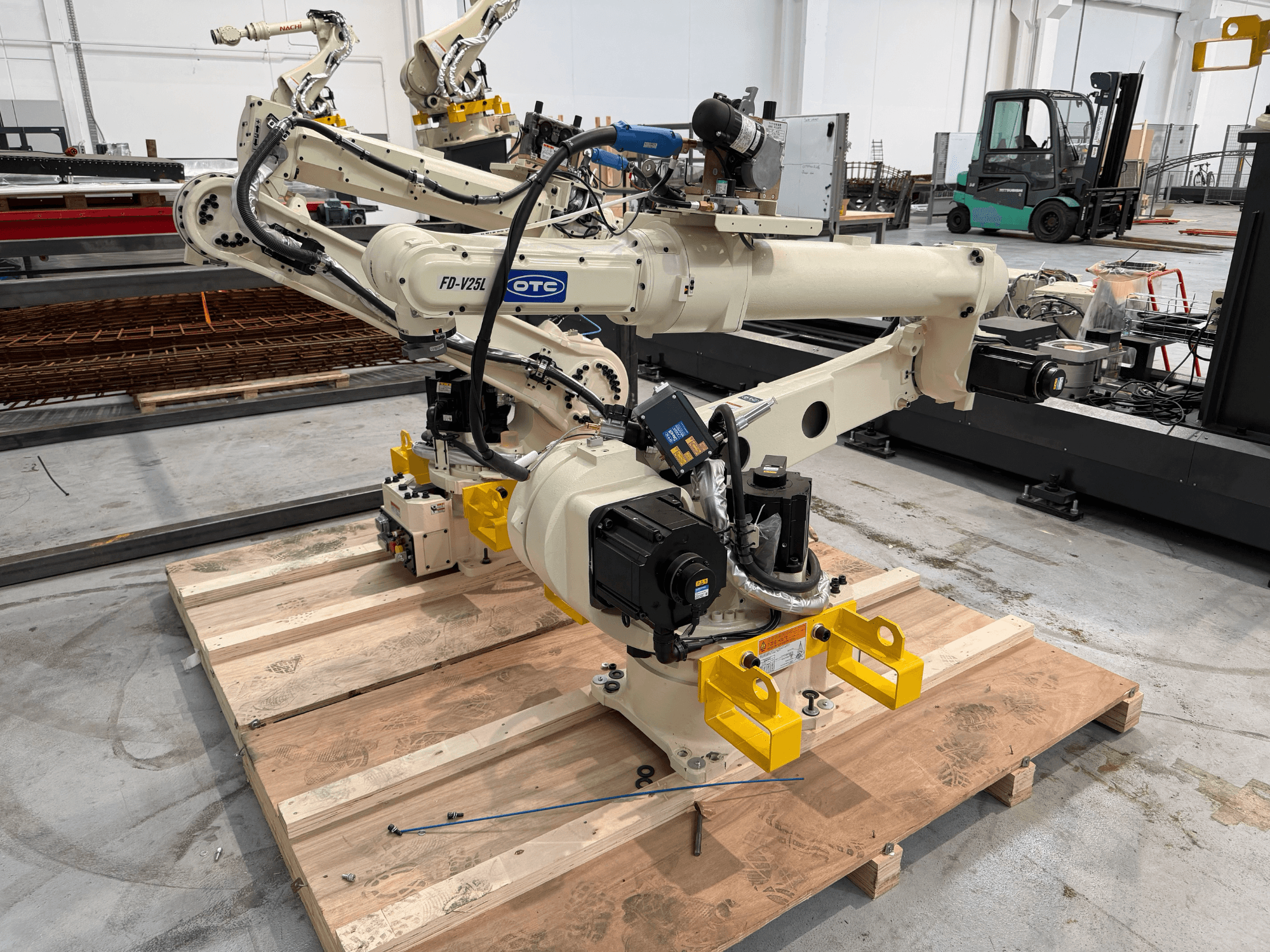Az OTC Daihen FD19-V25L robotkar elölnézetben, sárga fogókarokkal és látható mechanikus alkatrészekkel, egy fa raklapon.