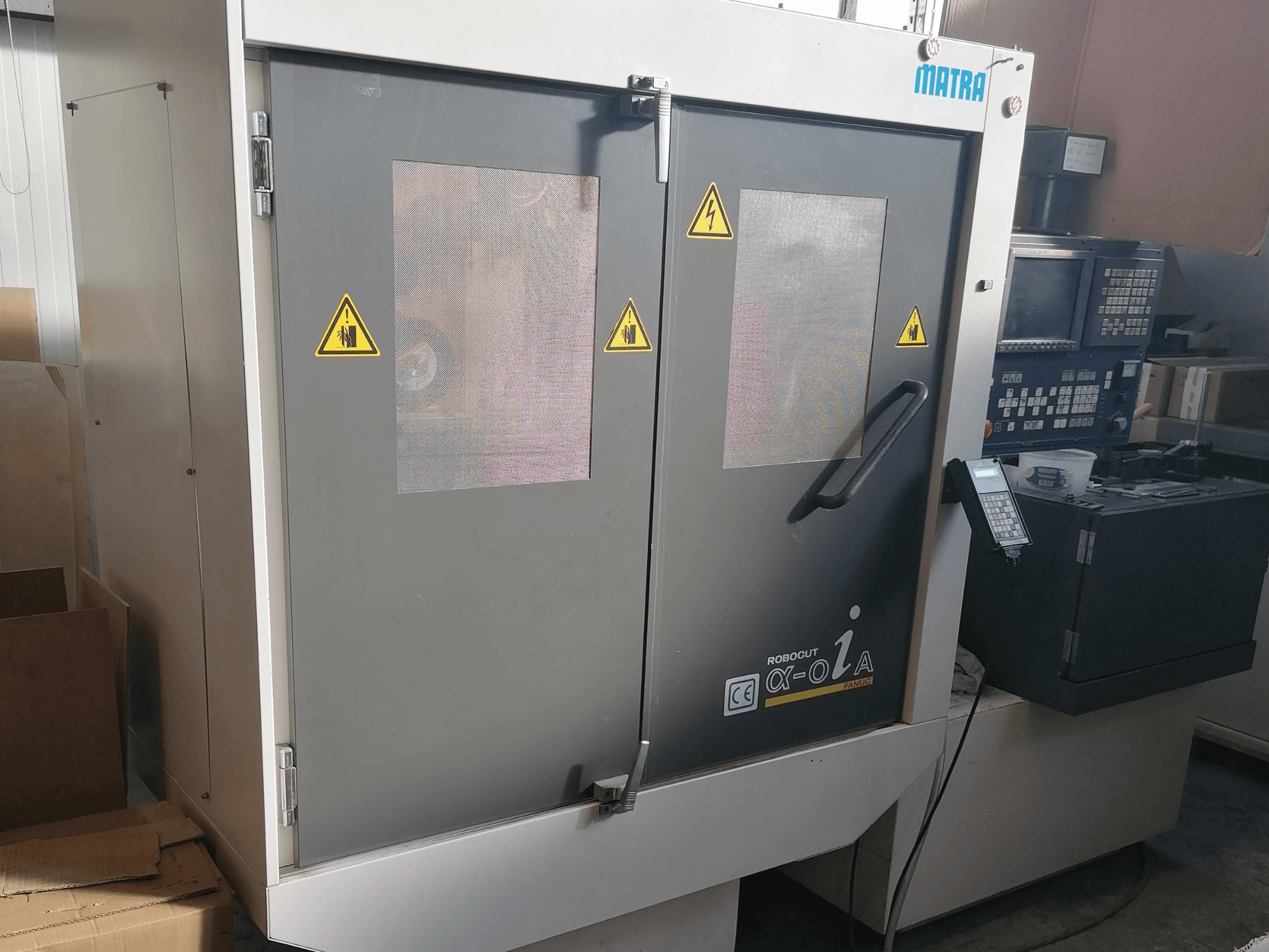 Robocut α-oZα gép a Matra-tól, elölnézetben a biztonsági figyelmeztetések, a vezérlőpanel és a hálós ablakokkal ellátott kettős ajtó.