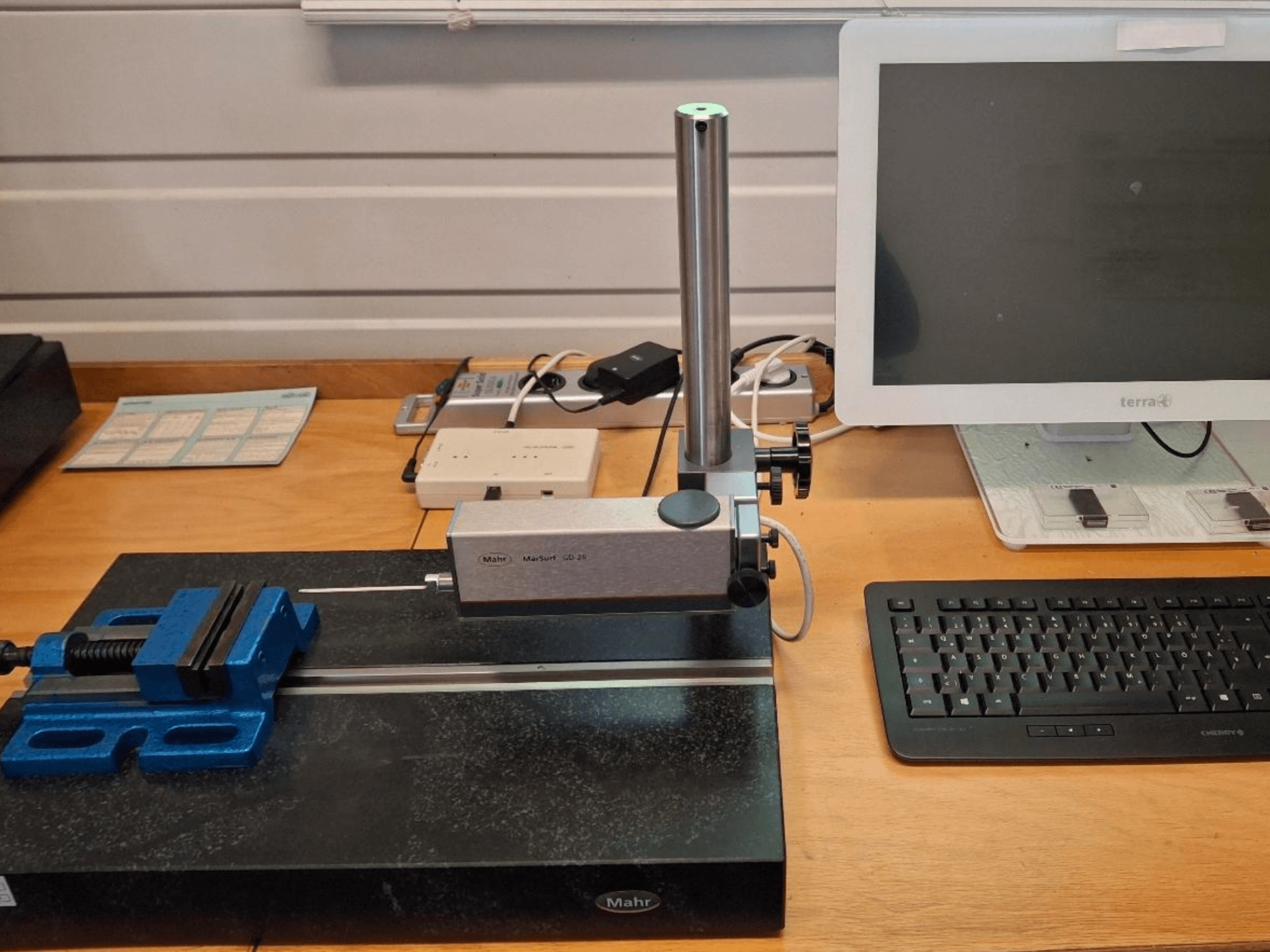 MAHR MARSURF GD 26 CMM egy munkapadon, mérőszondával, kék csavarhúzóval és számítógépes monitorral. Előlnézet.