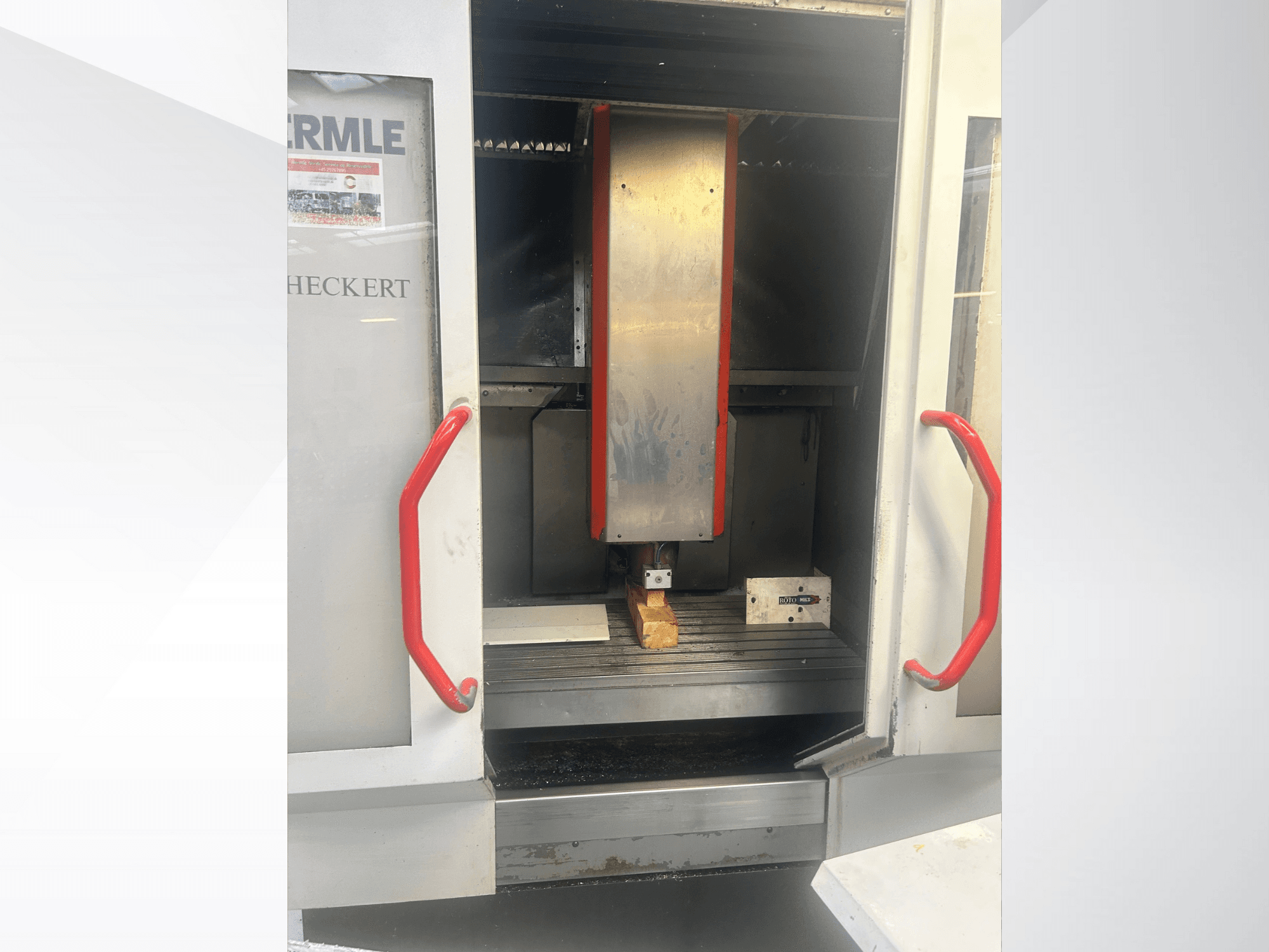 Hermle CNC-gép előlapja, fém belsővel, piros fogantyúkkal és egy munkadarabbal az asztalon.