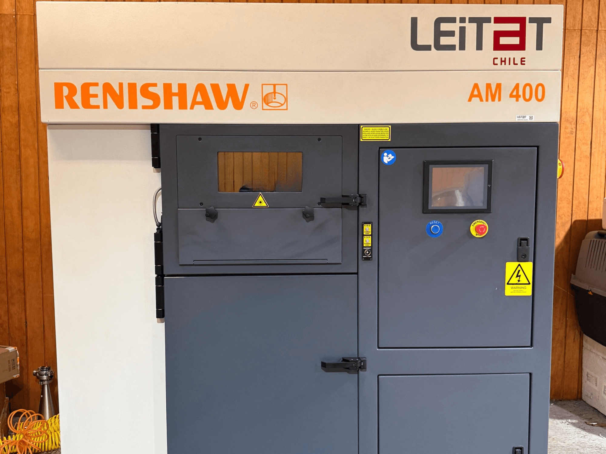 A RENISHAW AM400 fém 3D-nyomtató elölnézete, amelyen látható a vezérlőpanel, a figyelmeztető címkék és a karbantartási nyílás.