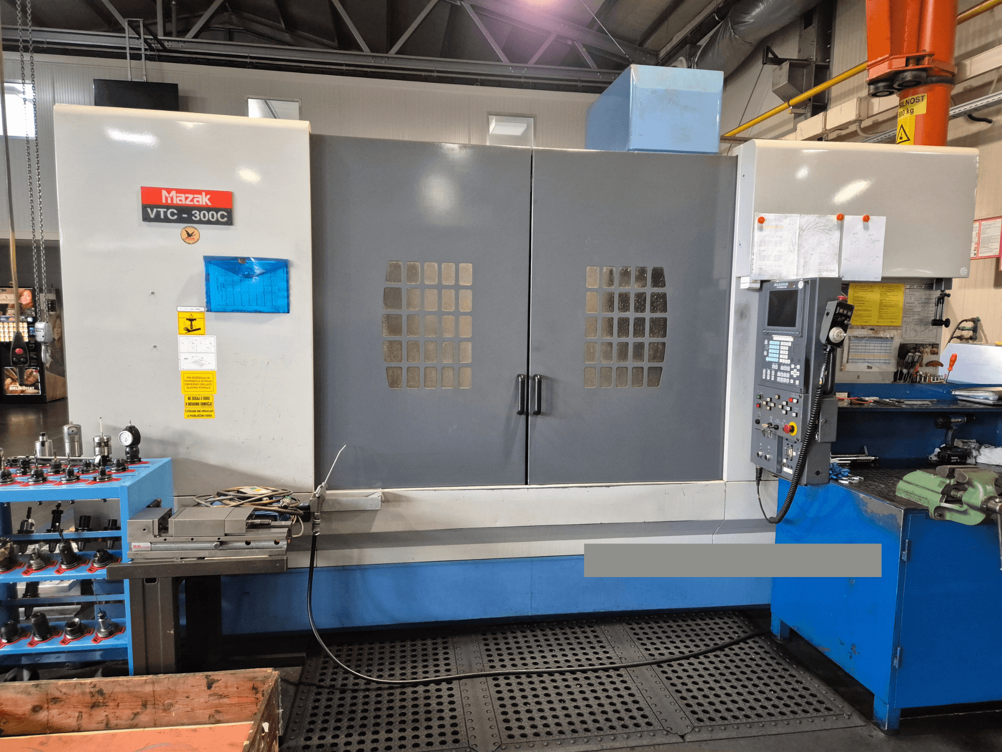 Mazak VTC-300C gép egy gyárban, elölnézetben a vezérlőpanel, a szürke ajtók ablakráccsal és a közeli berendezések.