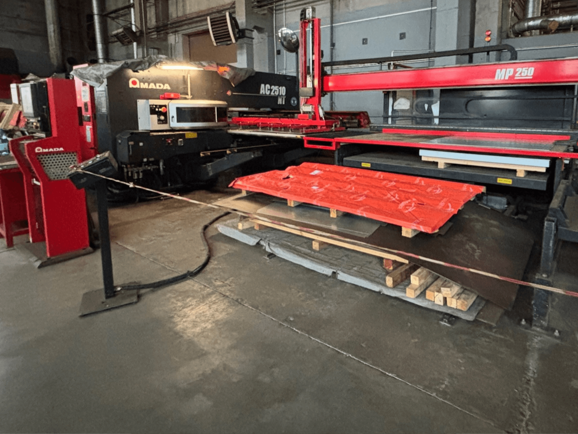 Amada AC2510 NT lézervágógép gyári környezetben, elölnézetben, nehézgépek, fémlemezek és raklapok a padlón.