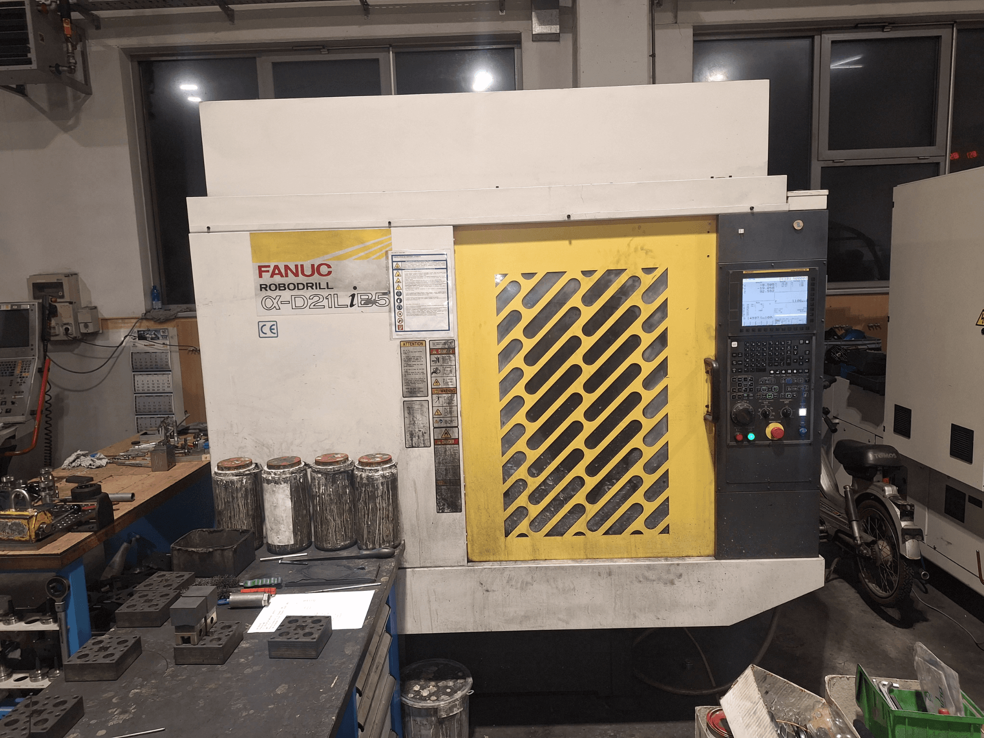 A FANUC Robodrill α-D21LiB5 előlapja, a kezelőpanel, a sárga rács és a közeli szerszámok a munkapadon.