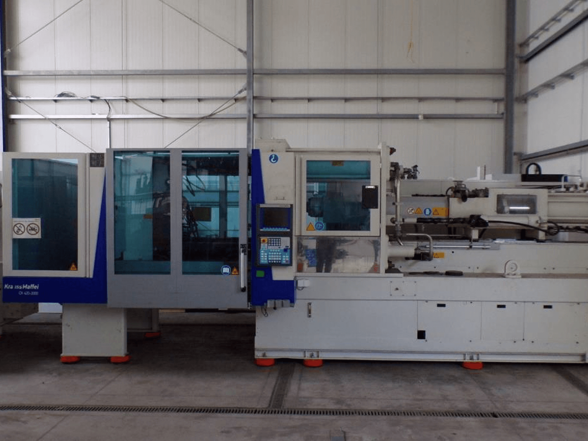 KraussMaffei fröccsöntőgép, oldalnézet, vezérlőpanel és biztonsági jelzők, látható gyári környezetben.