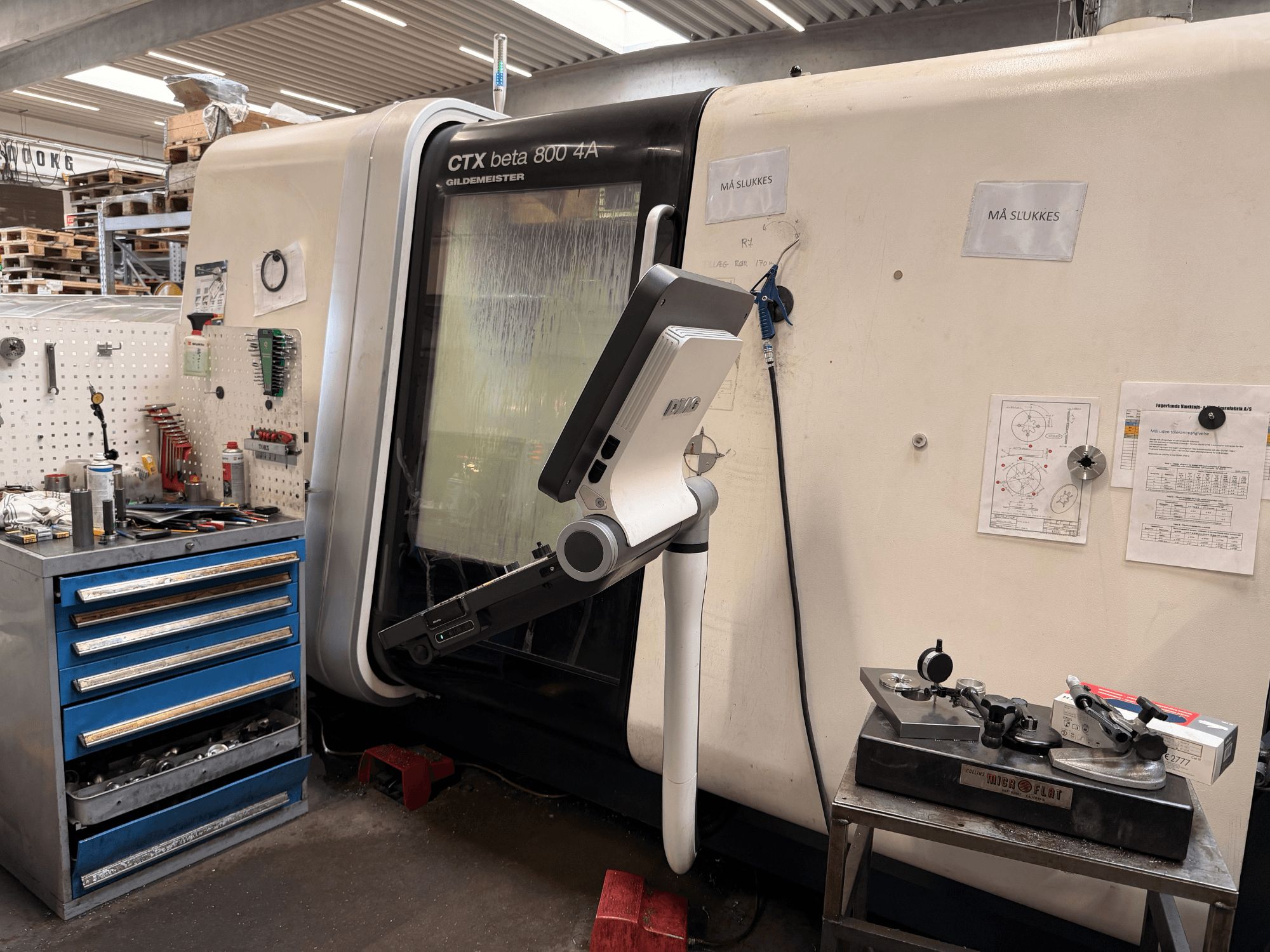 DMG MORI CTX Beta 800 4a függőleges esztergagép egy műhelyben, a közelben egy vezérlőkar és műszaki dokumentumok.