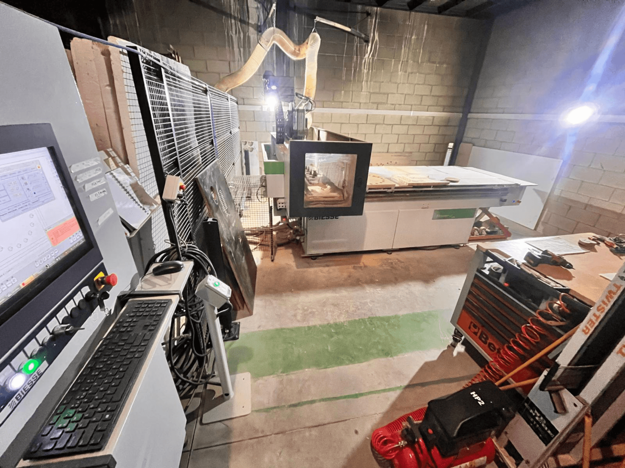 Biesse CNC-gép egy műhelyben, oldalnézetben a vezérlőpanel, a porelszívás, a szerszámok és a különböző anyagok az asztalon.