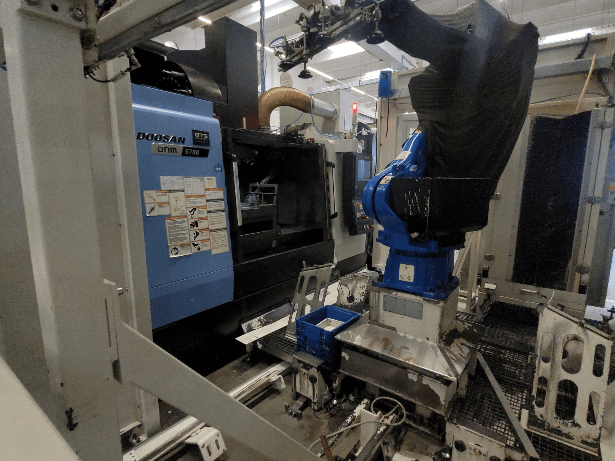 A DOOSAN DNM 5700 függőleges megmunkáló központ gyári környezetben, elölnézetben és a robotkarral való integrációját bemutatva.