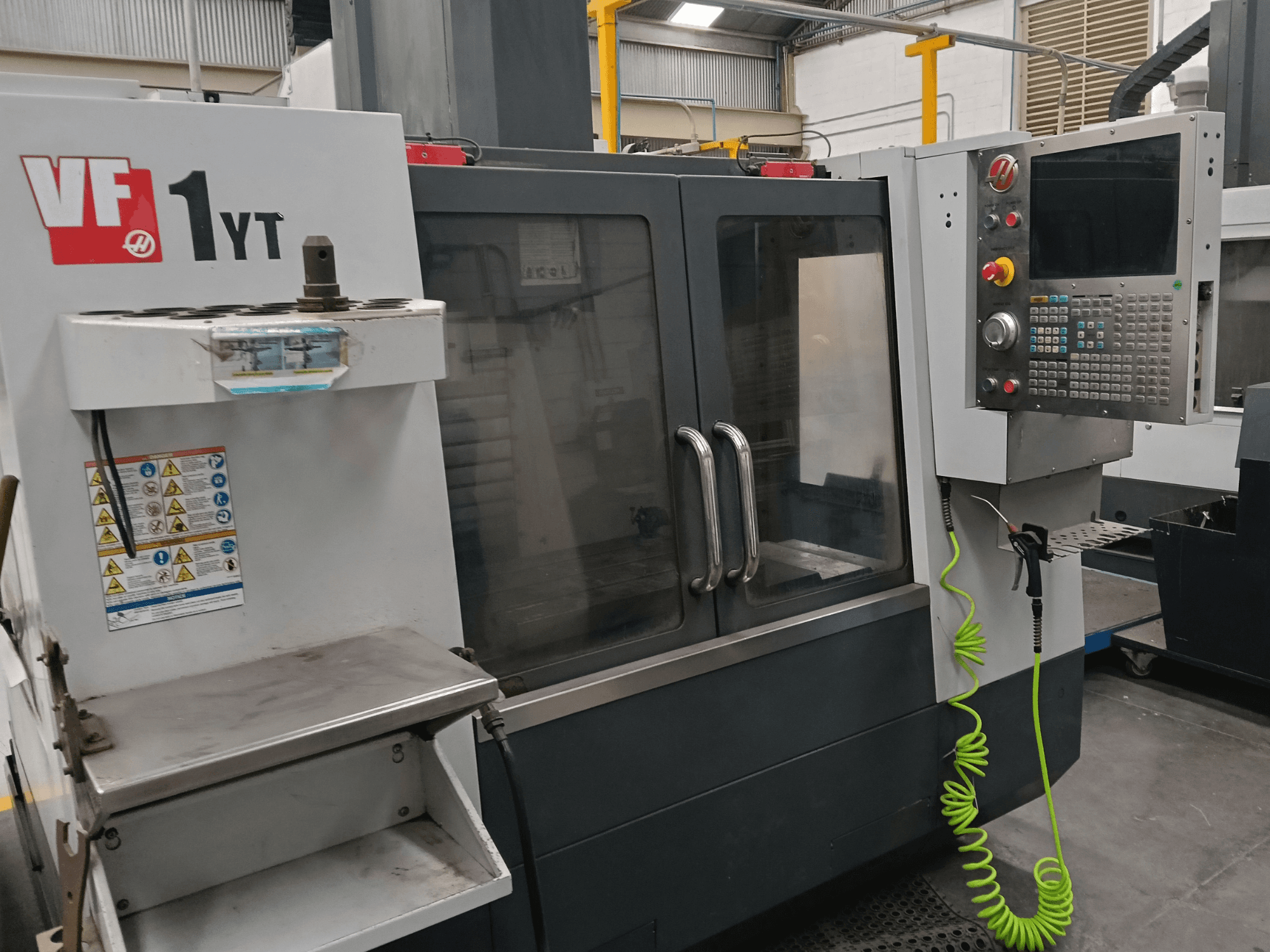 HAAS VF-1YT-2 függőleges megmunkáló központ, elölnézet, amelyen látható a vezérlőpanel, a szerszámtartó, a biztonsági címkék és a megmunkálási terület.