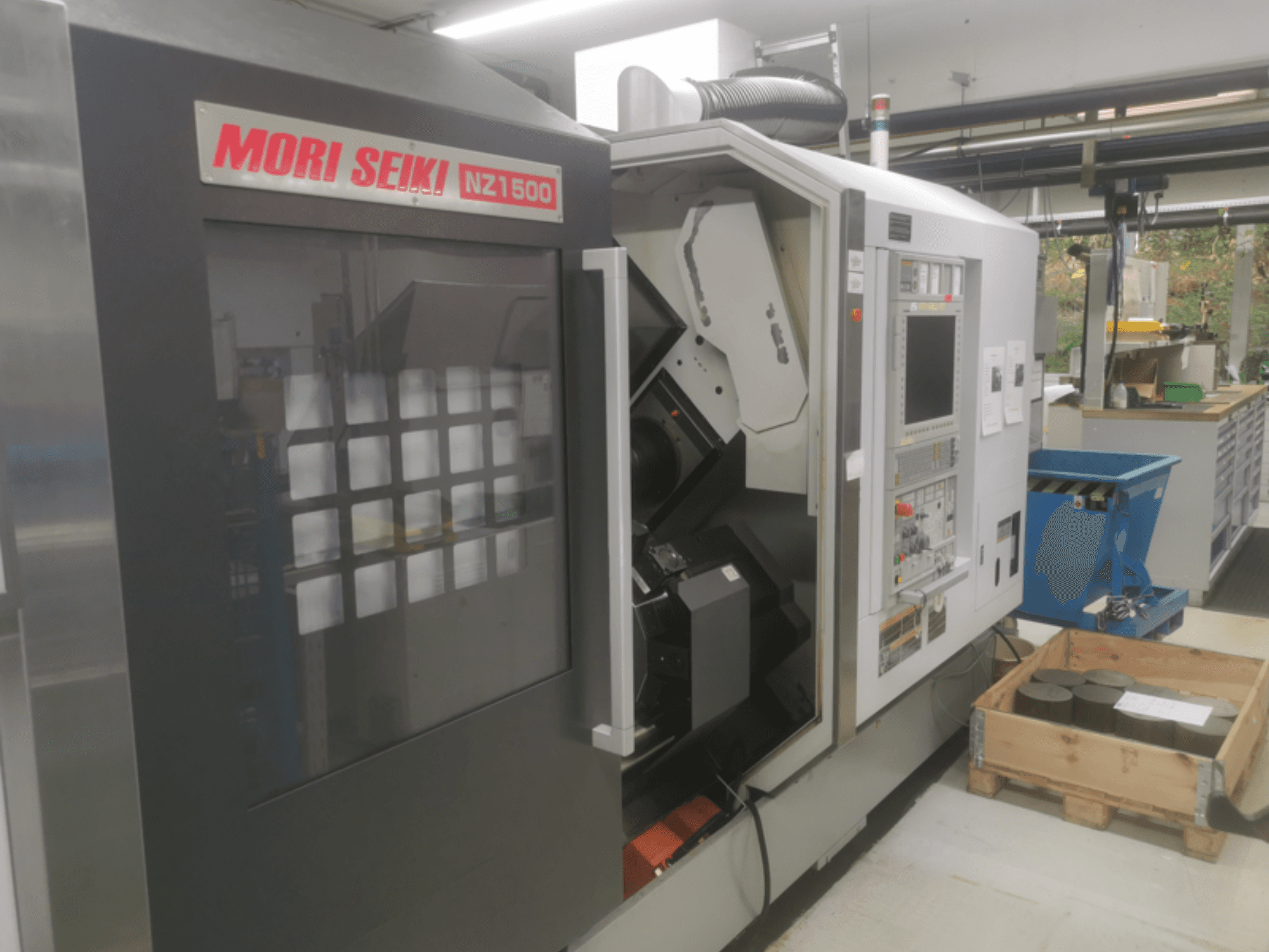MORI SEIKI NZ1500 CNC-gép enyhe szögben, a vezérlőpanel és a munkaterület látható a fapalettán lévő fém munkadarabokkal.