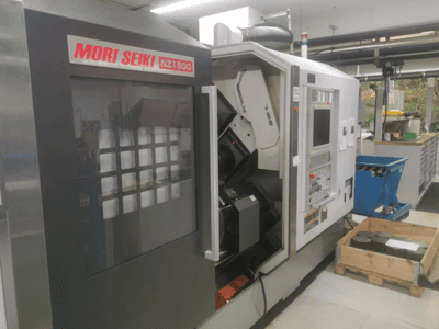 MORI SEIKI NZ1500 CNC-gép enyhe szögben, a vezérlőpanel és a munkaterület látható a fapalettán lévő fém munkadarabokkal.