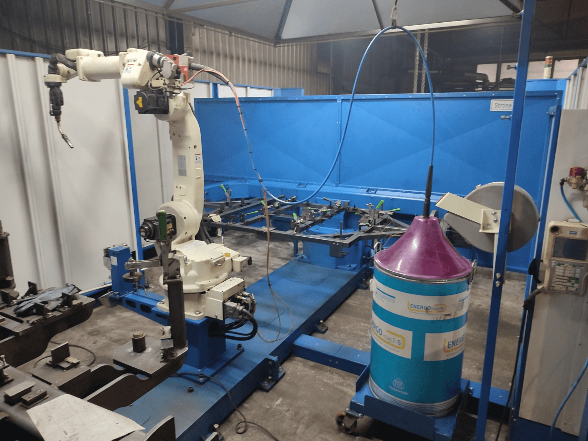 Ipari robotkar fehér és kék alapon, egy műhelyben, kék munkafelülettel és fém alkatrészekkel.